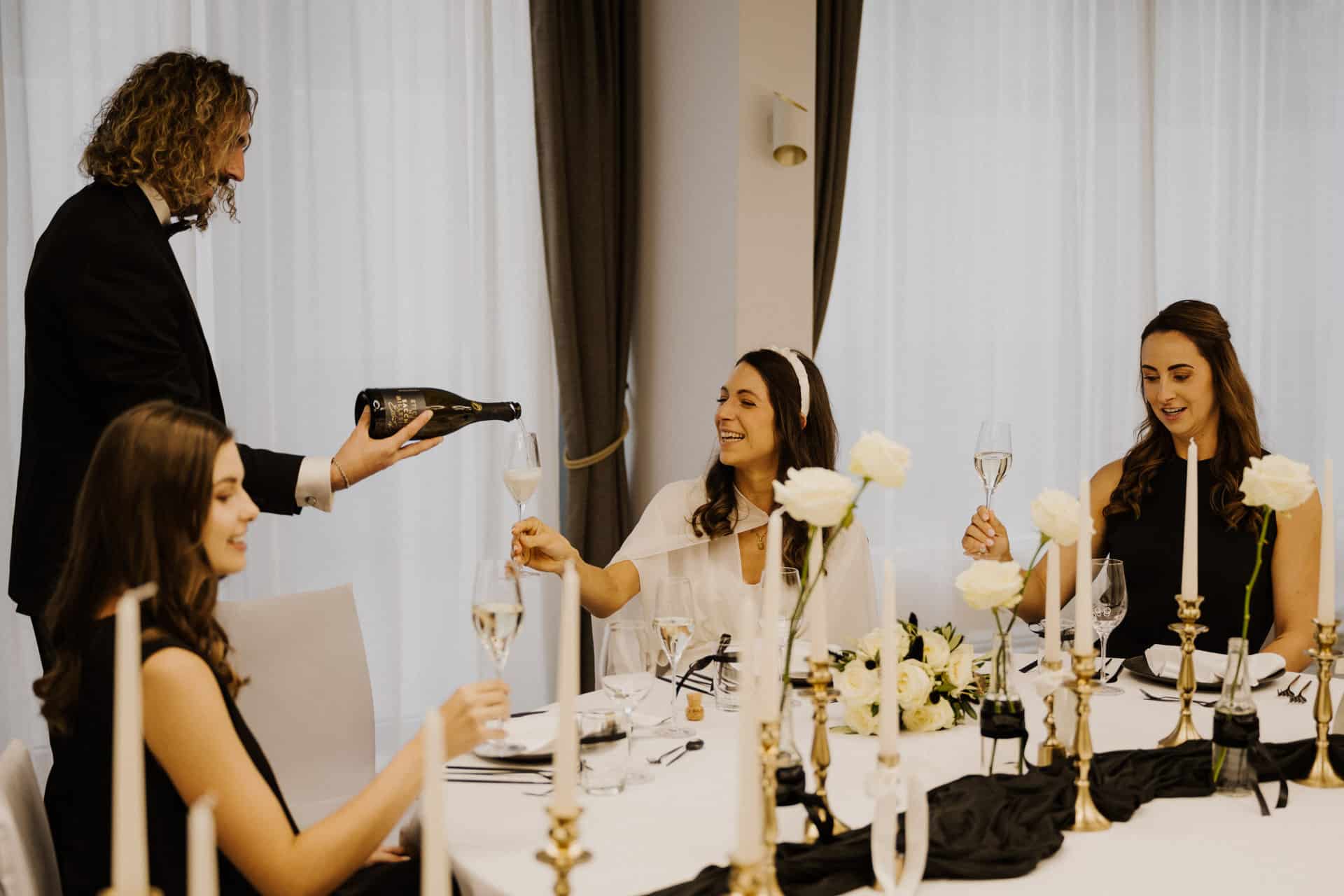 Ein Mann im Anzug schenkt drei lächelnden Frauen an einem eleganten, mit weißen Rosen, Kerzen und frischem Leinen geschmückten Lebensfeiertisch in einem hellen Raum mit großen Fenstern Champagner ein.