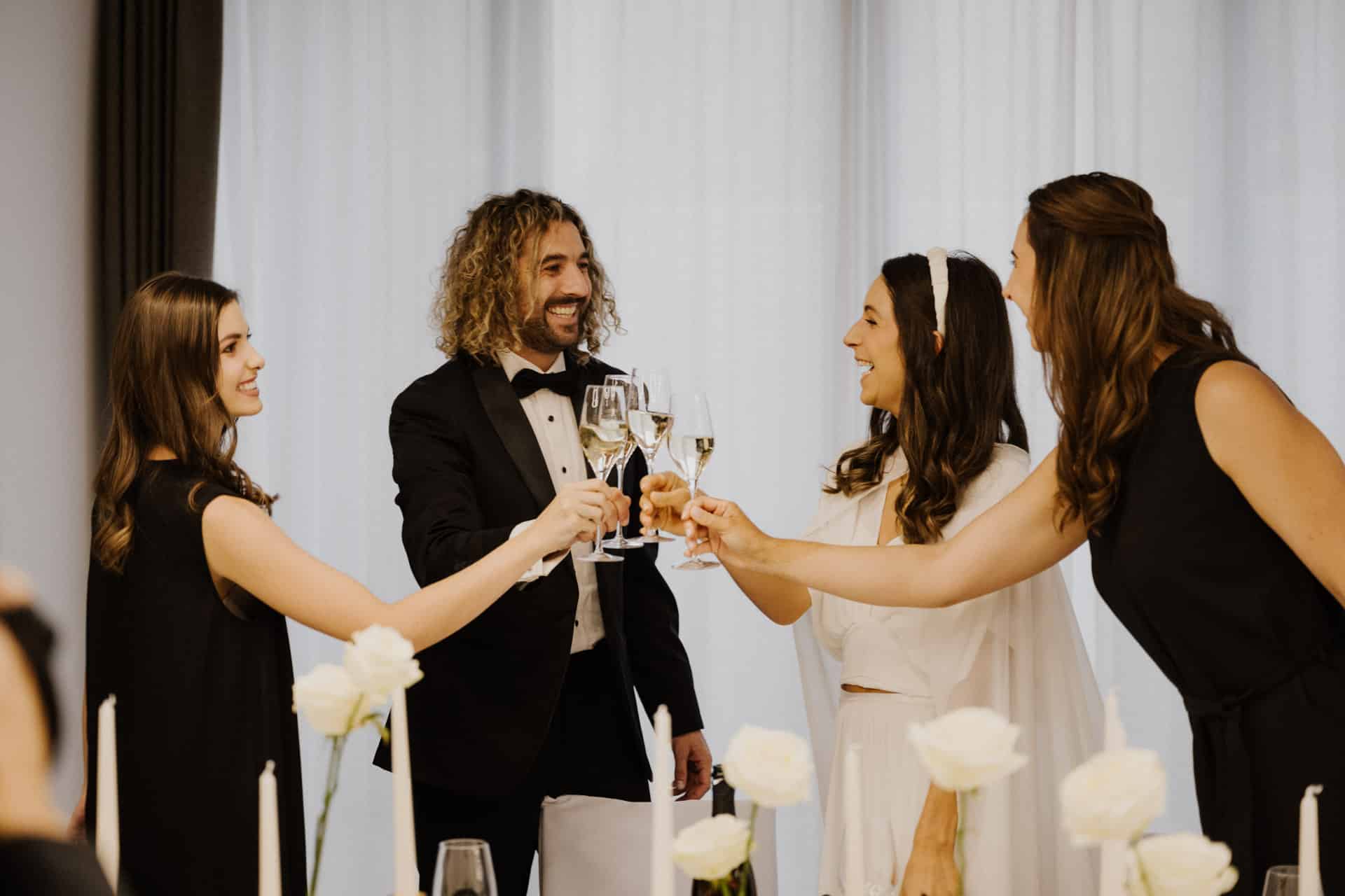 Vier förmlich gekleidete Personen, darunter eine Braut und ein Bräutigam, stoßen lächelnd mit Sektgläsern auf diese fröhlichen Lebensfeiern an. Weiße Blumen und Kerzen schmücken elegant den Tisch vor ihnen.