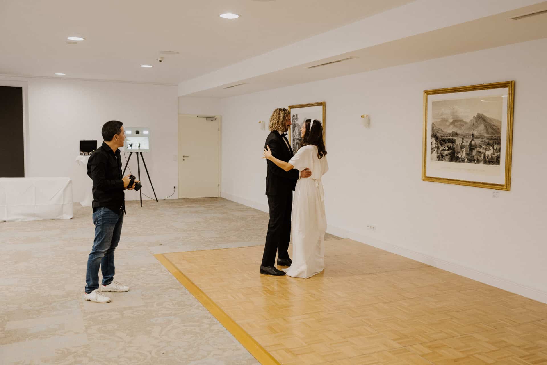 Ein Paar tanzt während der Lebensfeiern auf einer kleinen hölzernen Tanzfläche in einer Kunstgalerie, während ein Fotograf den Moment festhält. Der Raum ist mit hellen Wänden, gerahmten Kunstwerken und sanfter Beleuchtung ausgestattet.