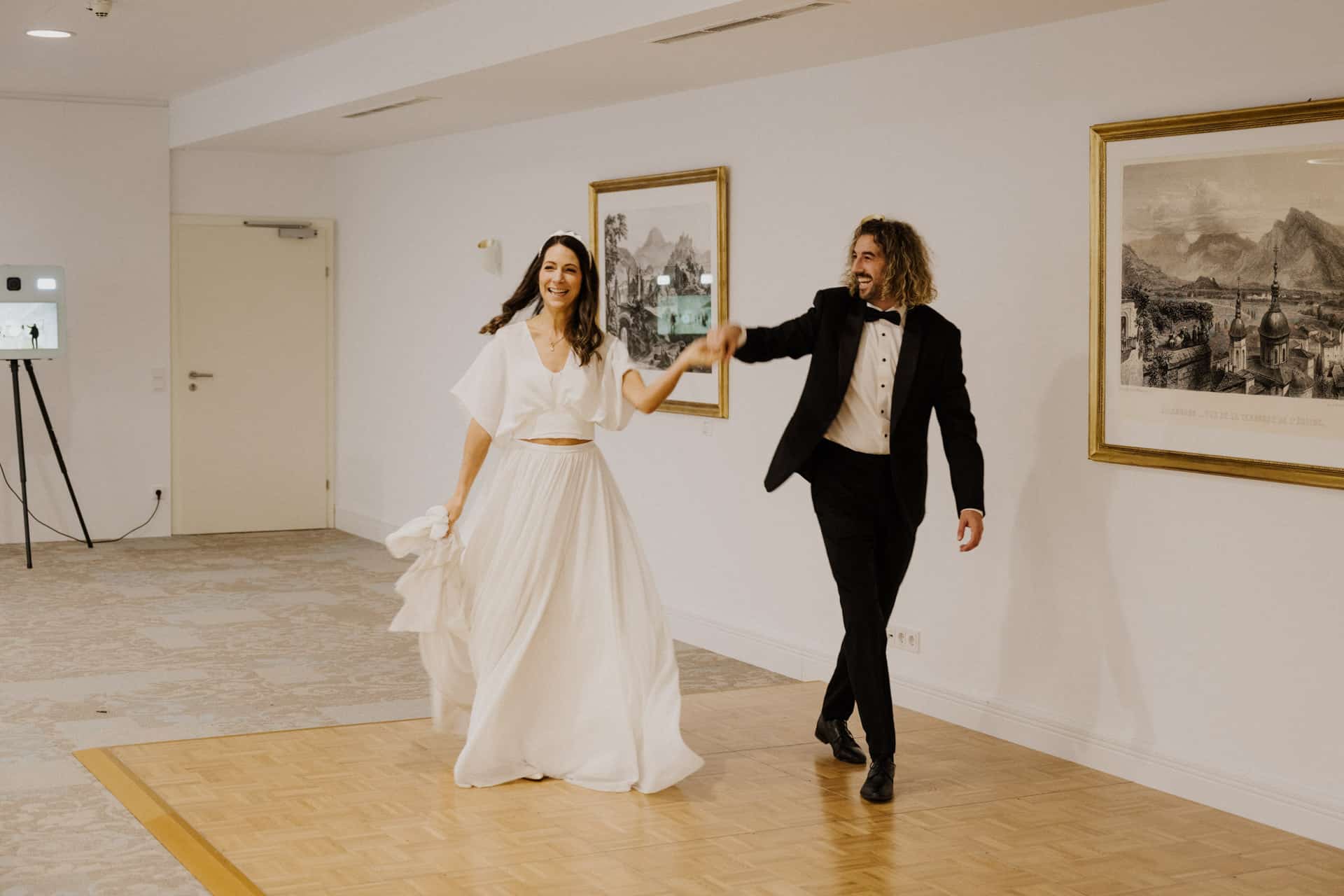 Eine Braut in einem weißen Kleid und ein Bräutigam in einem schwarzen Smoking halten sich an den Händen und lächeln, während sie gemeinsam auf einem Holzboden in einem hellen, eleganten Raum mit gerahmter Kunst tanzen - ein echter Lebensfeiern-Moment.