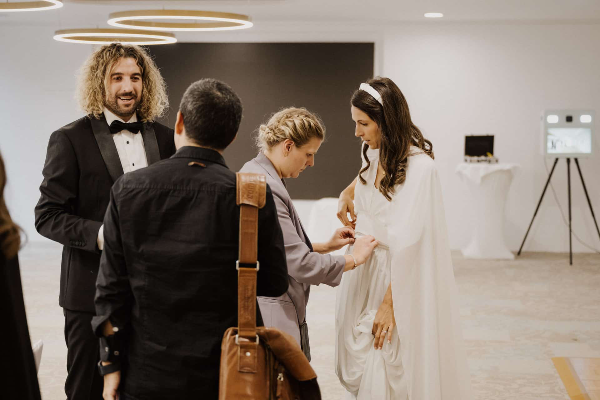 Eine Braut in einem weißen Kleid wird von einer Frau bei ihrem Outfit unterstützt, während ein Mann im Smoking und ein weiterer Mann mit einer ledernen Umhängetasche in der Nähe stehen - alle versammeln sich zu Lebensfeiern in einem modernen, gut beleuchteten Raum.