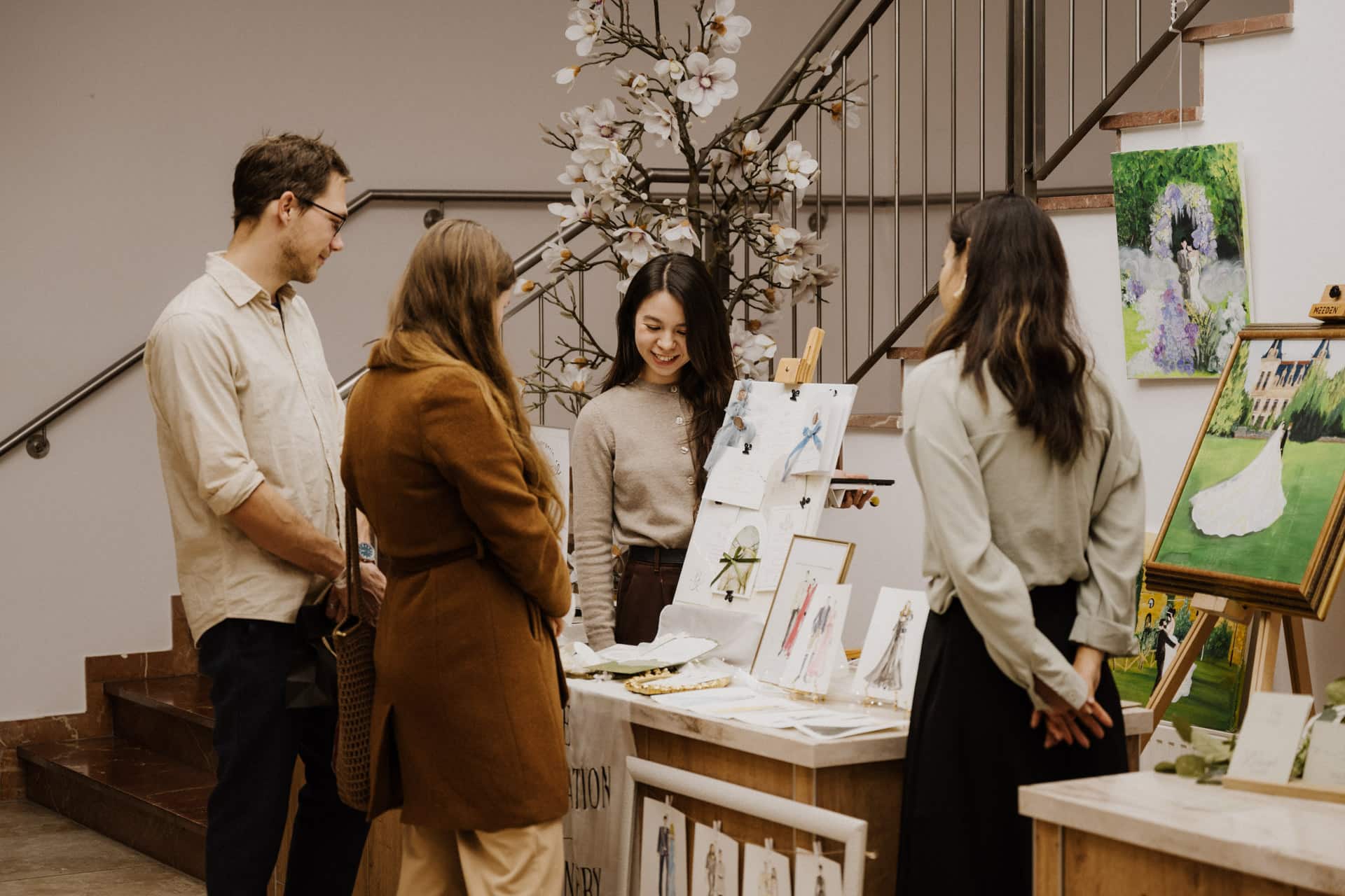 Vier Personen stehen um einen Tisch herum und stellen Kunstdrucke und Karten für Lebensfeiern aus. Eine Frau hinter dem Tisch lächelt, während sie sich mit den anderen unterhält. Kunstwerke und Gemälde sind auf dem Tisch und den Staffeleien in der Nähe angeordnet.