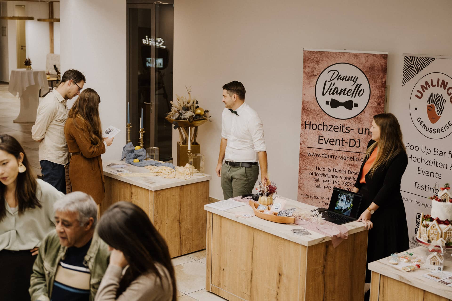 Bei einer Indoor-Veranstaltung mit Verkaufsständen interagieren die Menschen. Ein Stand wirbt für einen Hochzeits-, Event- und Lebensfeiern-DJ, ein anderer bietet Verkostungserlebnisse an. Auf den Tischen liegen Werbeartikel, Broschüren und festliche Dekorationen aus.