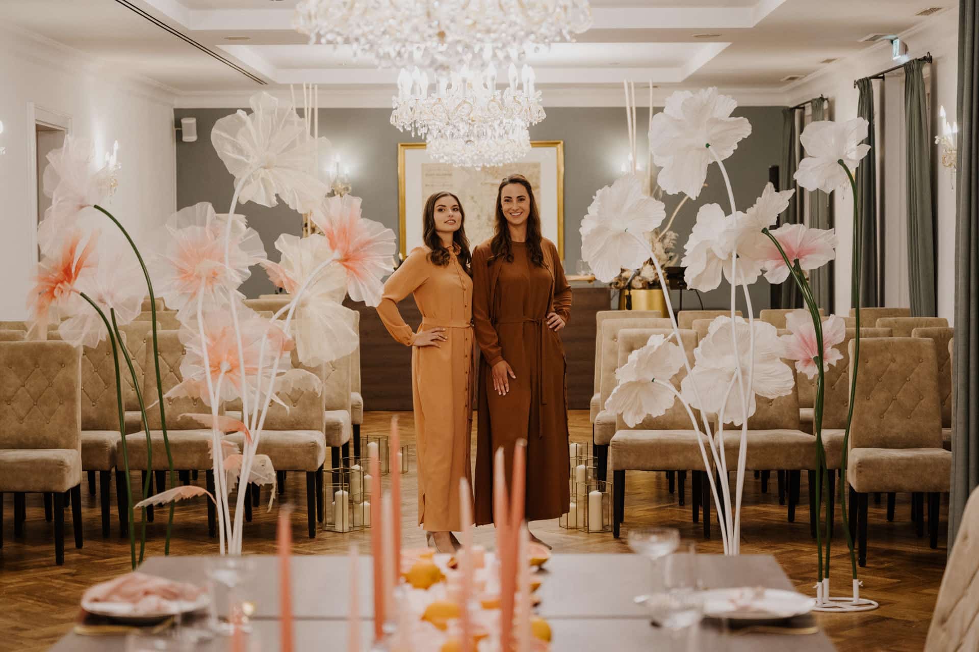 Zwei Frauen in langen braunen Kleidern stehen Seite an Seite in einem eleganten, geschmückten Raum. Große weiße und rosafarbene Blumenarrangements säumen den Gang für die Lebensfeiern, und über den Reihen brauner Stühle leuchten Kronleuchter.