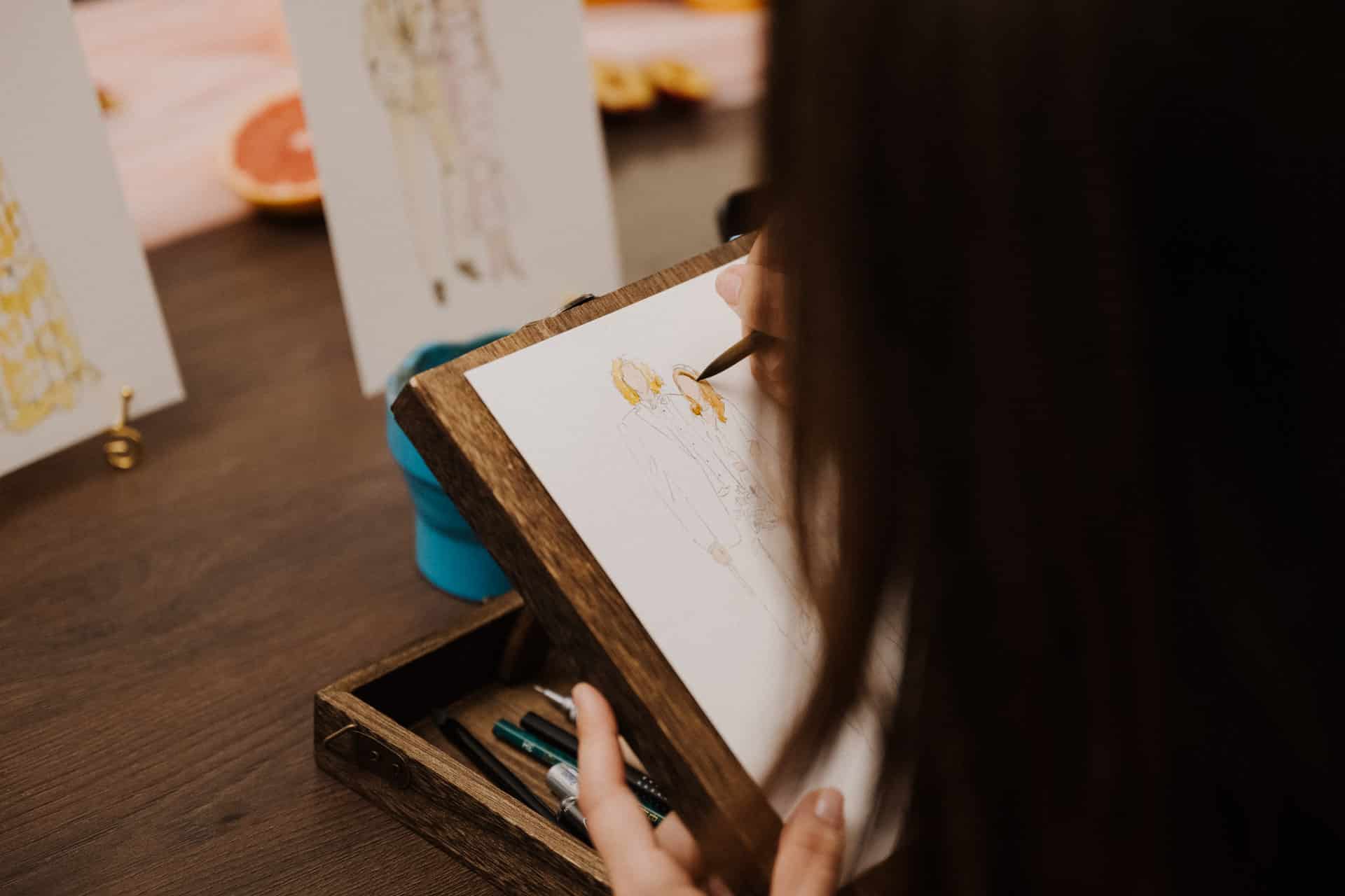 Eine Person skizziert mit Farbstiften eine Modeillustration auf einem Holztisch, umgeben von Zeichengeräten und anderen Skizzen, und feiert Lebensfeiern mit einem Kunstwerk, das eine Figur in leuchtenden Gelbtönen zeigt.