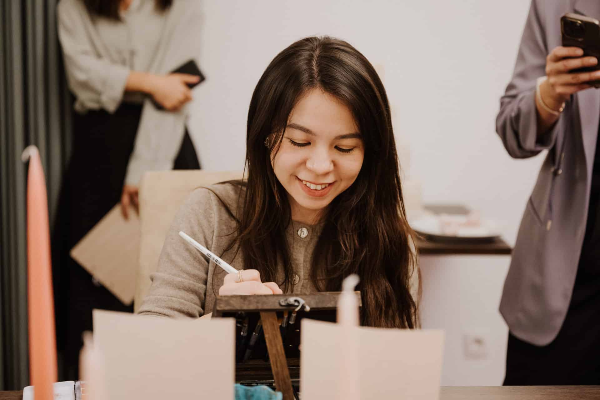 Eine junge Frau mit langen dunklen Haaren lächelt, während sie während eines Lebensfeiern-Treffens an einem Schreibtisch schreibt. Zwei Personen stehen in der Nähe, eine hält ein Notizbuch in der Hand, die andere telefoniert. Auf dem Tisch liegen verschiedene Papiere und Kerzen.
