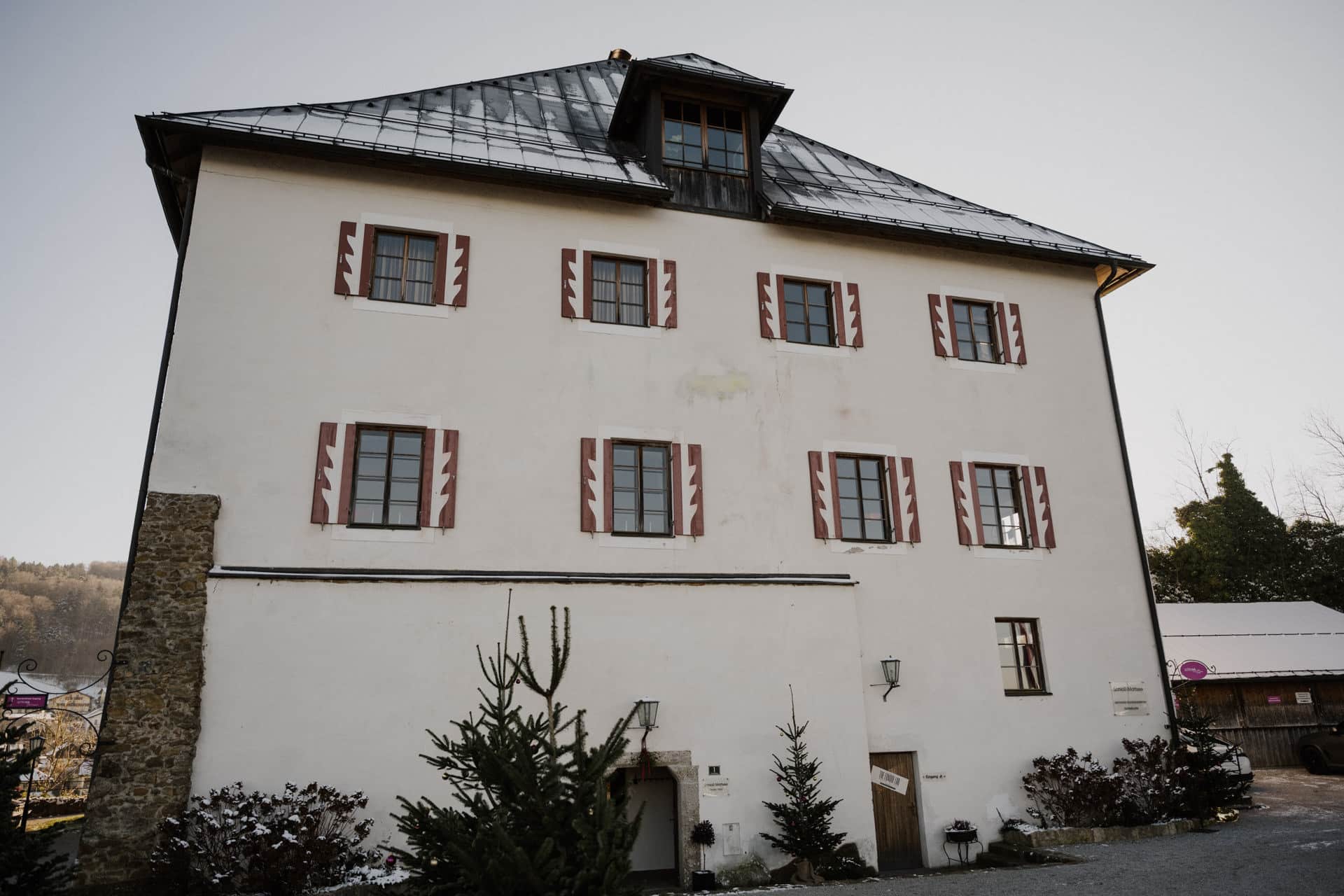 Ein dreistöckiges weißes Gebäude mit einem dunklen Metalldach, roten und weißen Fensterläden und einem steinernen Eckpfeiler. Davor sind kleine Bäume und Sträucher gepflanzt, und der Himmel ist klar.