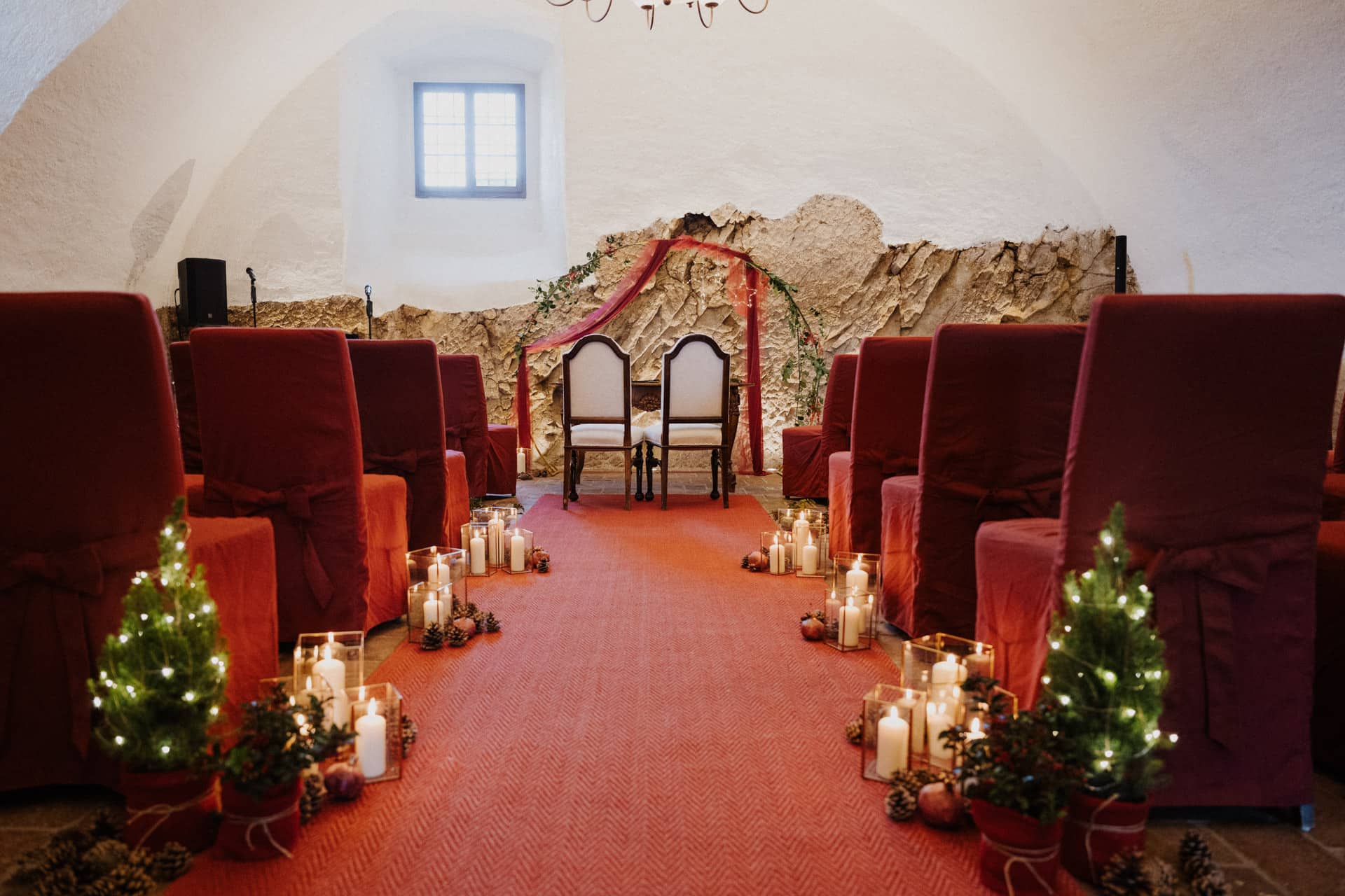Bei einer kleinen Hochzeitszeremonie stehen zwei Stühle im Vordergrund, die von rot bezogenen Stühlen in Reihen flankiert werden. Der Gang ist mit Kerzen, Tannenzapfen, Grünzeug und kleinen beleuchteten Weihnachtsbäumen gesäumt, was eine festliche, gemütliche Atmosphäre schafft.