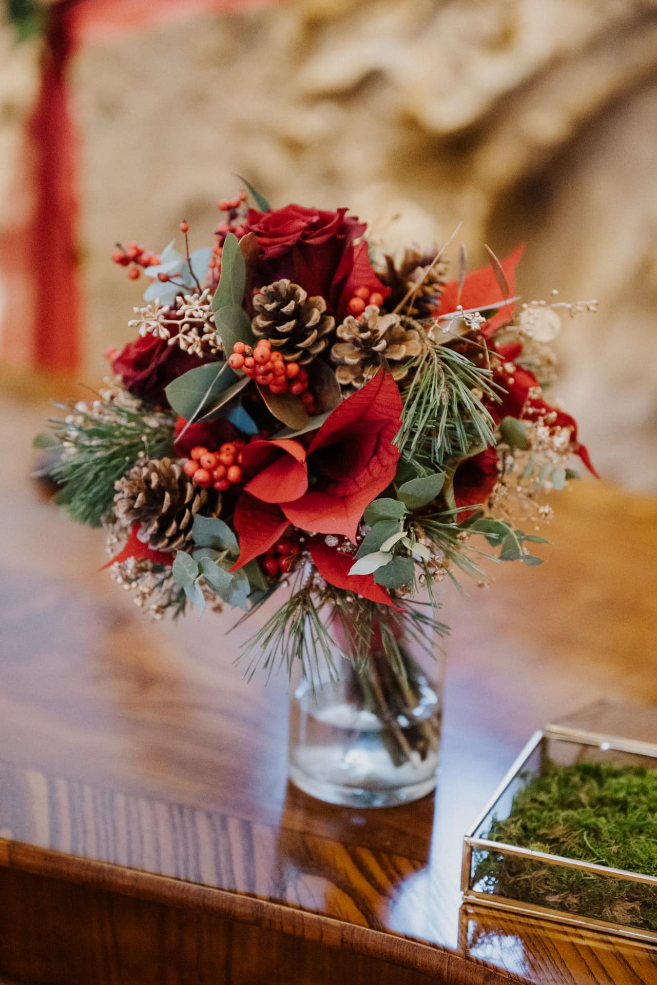 Ein festlicher Strauß in einer Glasvase mit roten Rosen, Tannenzapfen, roten Beeren, Eukalyptusblättern und grünen Tannennadeln, arrangiert auf einer Holzunterlage.