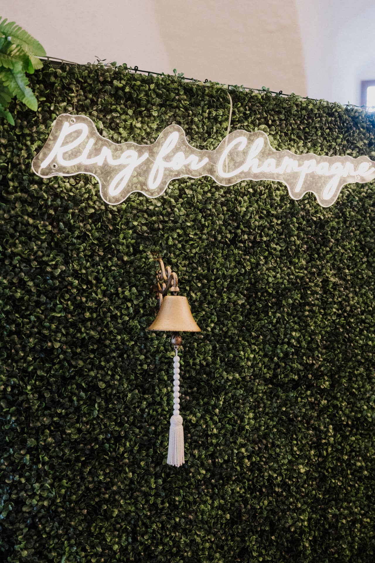 Eine Messingglocke mit einem weißen Perlengriff hängt an einer grünen, begrünten Wand. Darüber prangt ein Neonschild mit der Aufschrift "Ring for Champagne" in kursiven Buchstaben.