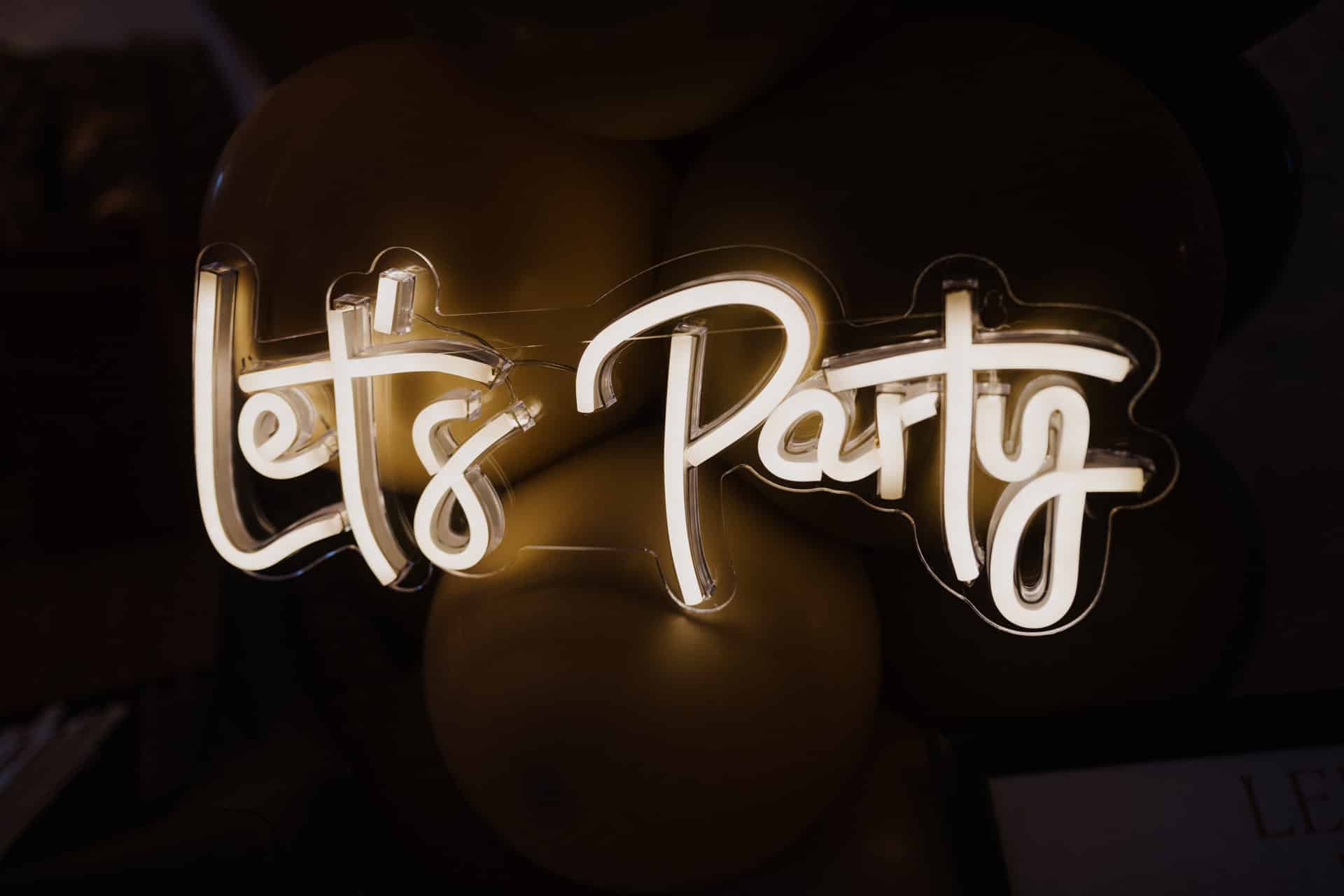 Ein leuchtendes Neonschild in Schreibschrift mit der Aufschrift "Let's Party" leuchtet vor einem dunklen Hintergrund mit schwach sichtbaren Luftballons dahinter.