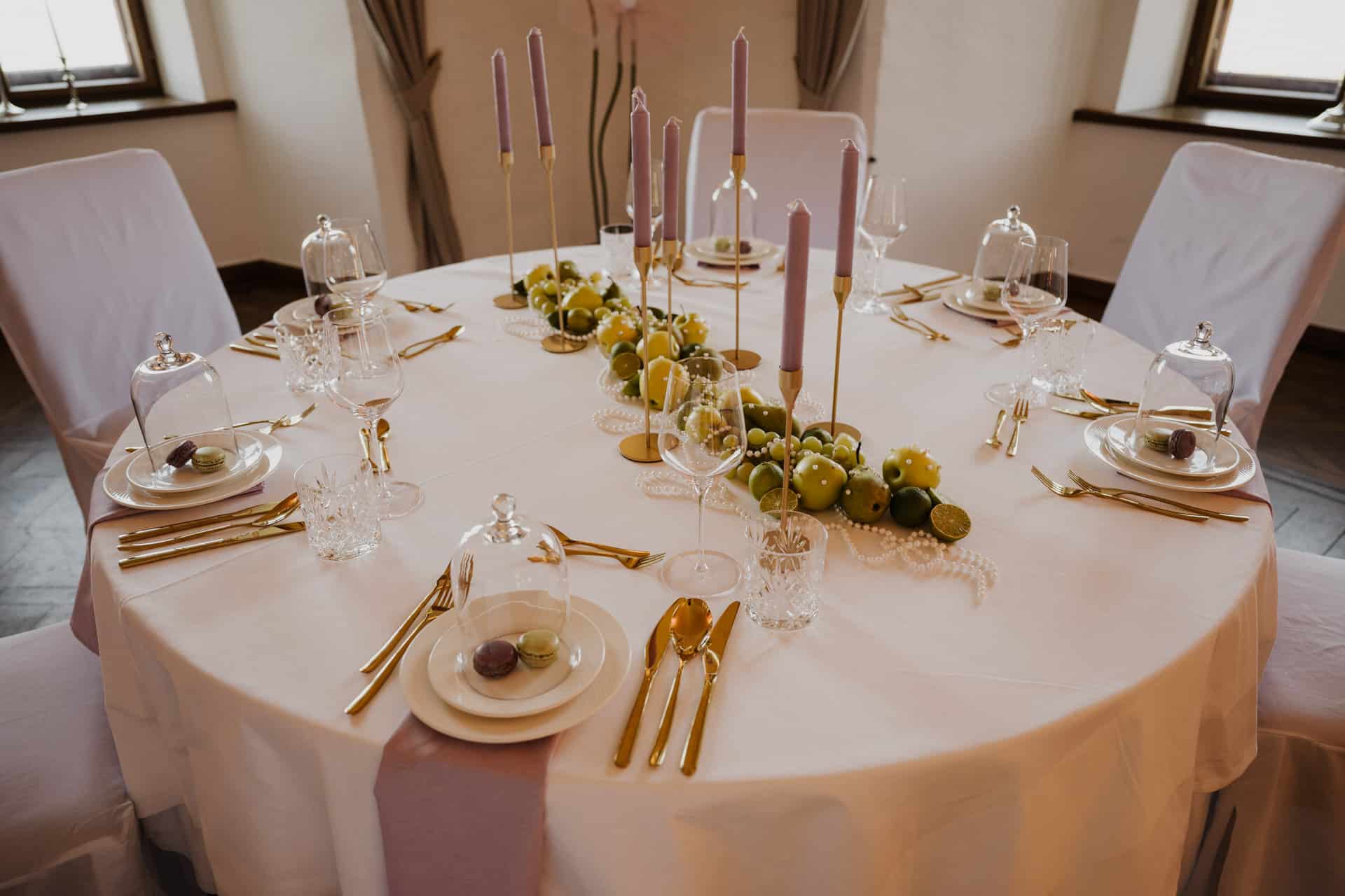 Ein runder Tisch für sechs Personen mit weißer Tischdecke, goldenem Besteck, Lavendelkerzen, grünen Äpfeln als Tafelaufsatz und Glaskuppeln, die Macarons auf jedem Teller bedecken, in einem eleganten, sanft beleuchteten Raum.