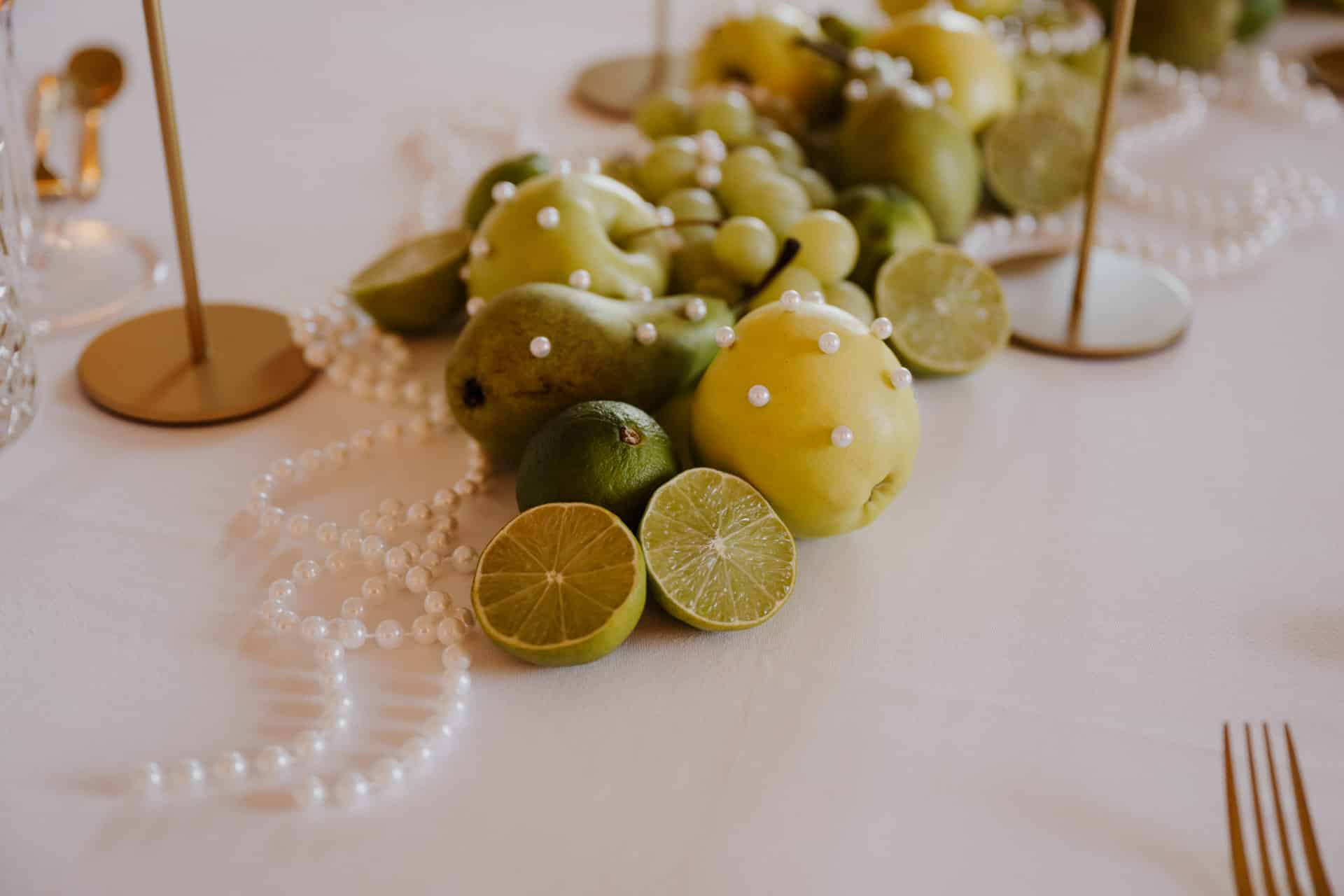 Ein Tafelaufsatz mit grünen Früchten wie Äpfeln, Limetten, Birnen und Trauben, verziert mit Perlensträngen, steht auf einem weißen Tischtuch, daneben stehen goldene Kerzenständer.