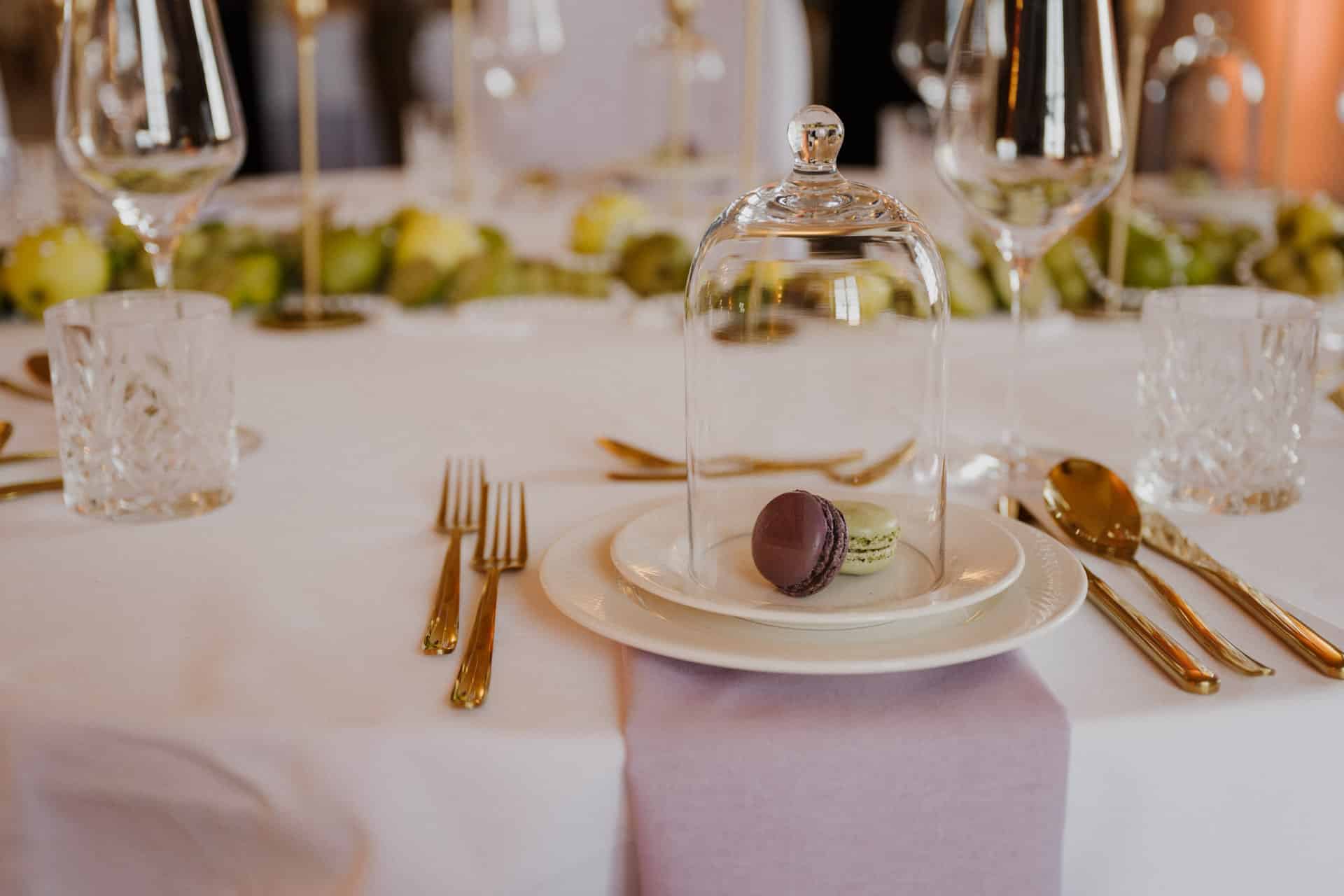 Ein gedeckter Tisch mit zwei Macarons unter einer Glasglocke auf einem weißen Teller, umgeben von goldenem Besteck, Kristallgläsern, Weingläsern und einer über den Rand drapierten lavendelfarbenen Serviette. Grüne Dekorationen verlaufen in der Mitte.