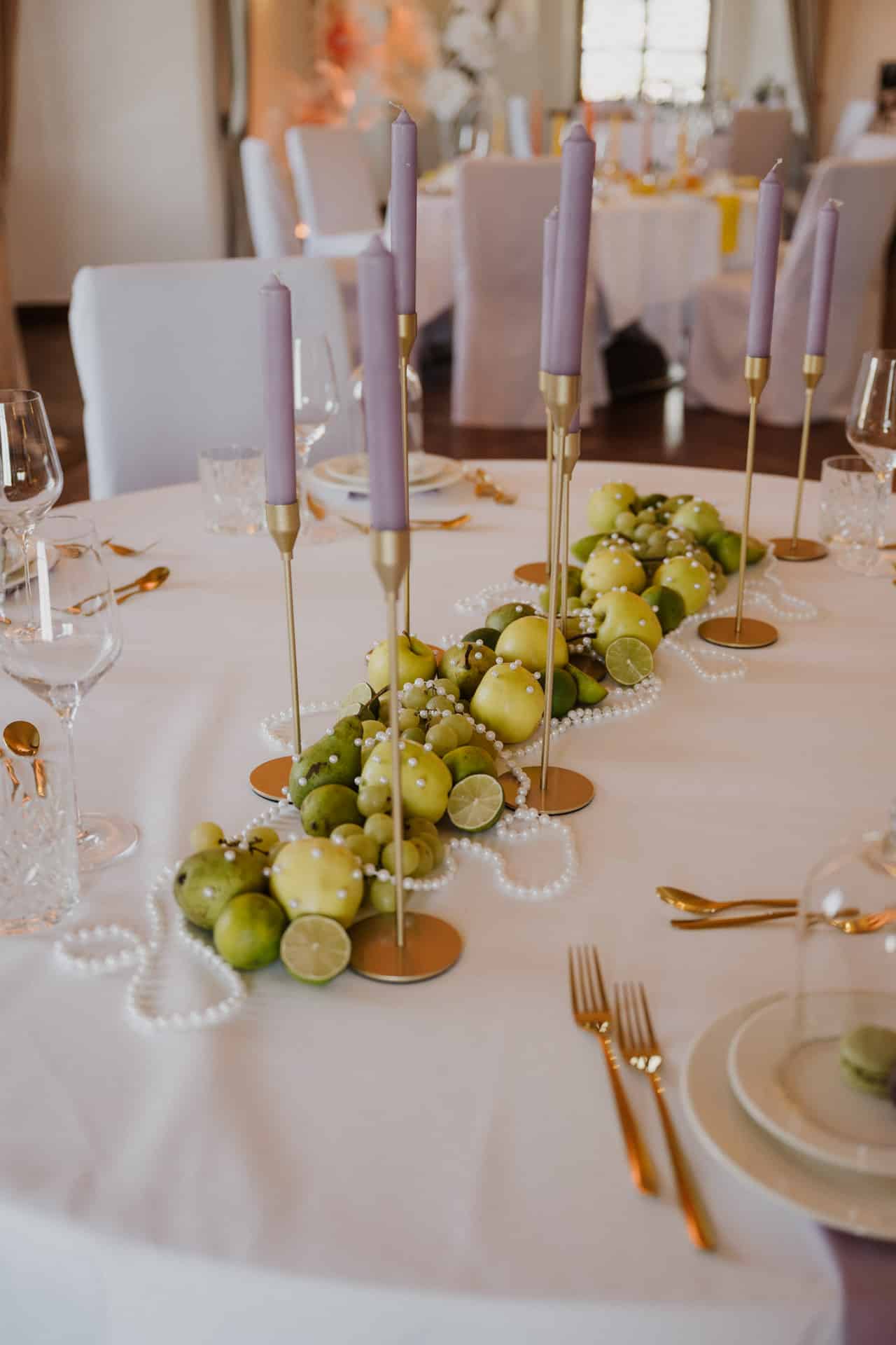 Ein runder Tisch mit weißem Tischtuch ist mit grünen Äpfeln, Limetten, weißen Perlen und goldenen Kerzenhaltern mit hohen fliederfarbenen Kerzen dekoriert. Elegante Gläser und goldenes Besteck sind auf dem Tisch angeordnet.