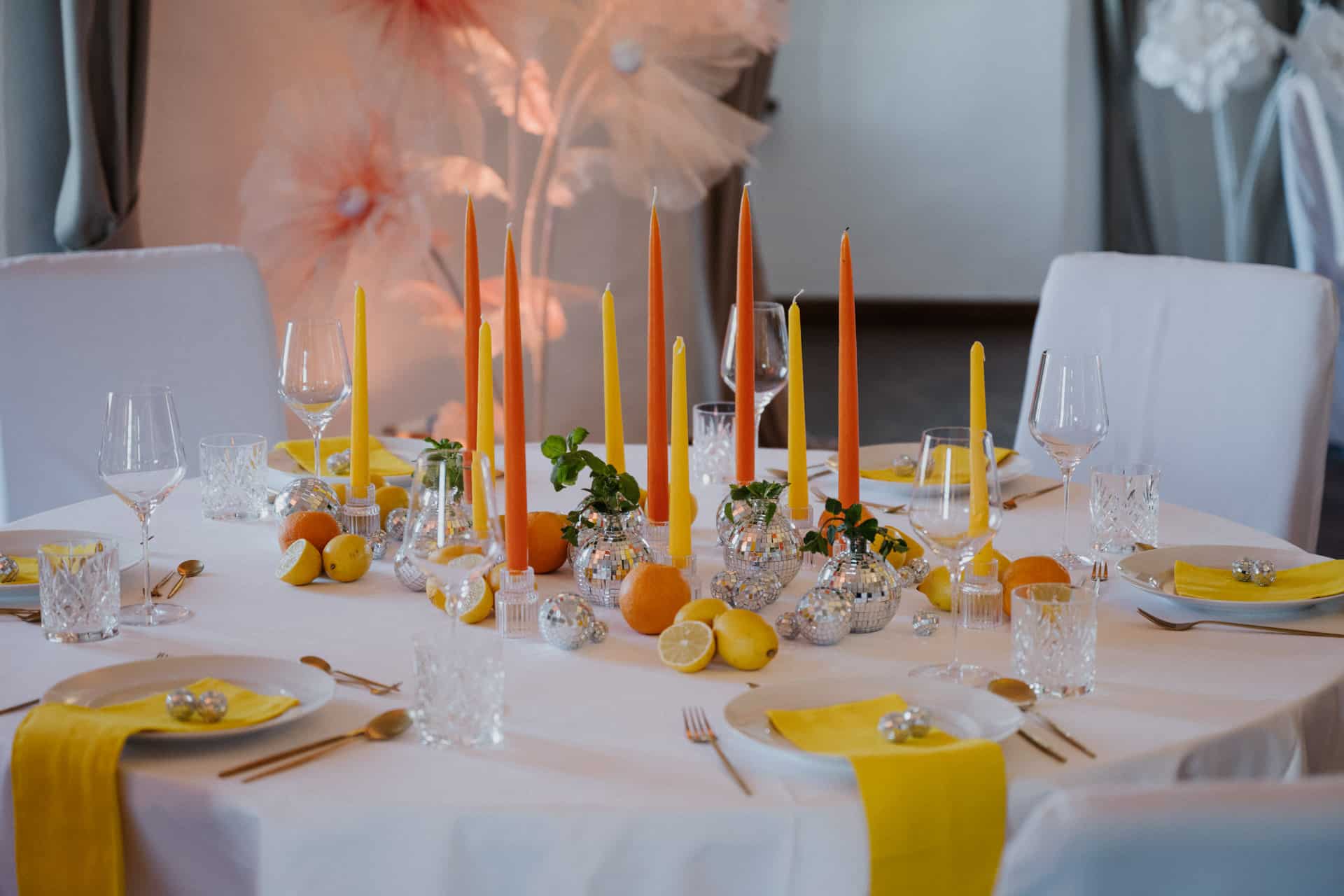 Ein runder Tisch für ein formelles Essen mit gelben und orangefarbenen Kerzen, Zitrusfrüchten, Glaswaren und weißen Tellern mit gelben Servietten schafft eine helle, festliche Atmosphäre.