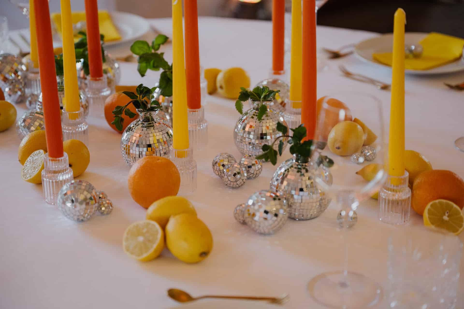 Eine Tischdekoration mit gelben und orangefarbenen Kerzen, Discokugel-Ornamenten, frischen Zitronen und Orangen auf einer weißen Tischdecke, die ein helles und festliches Ambiente schafft.