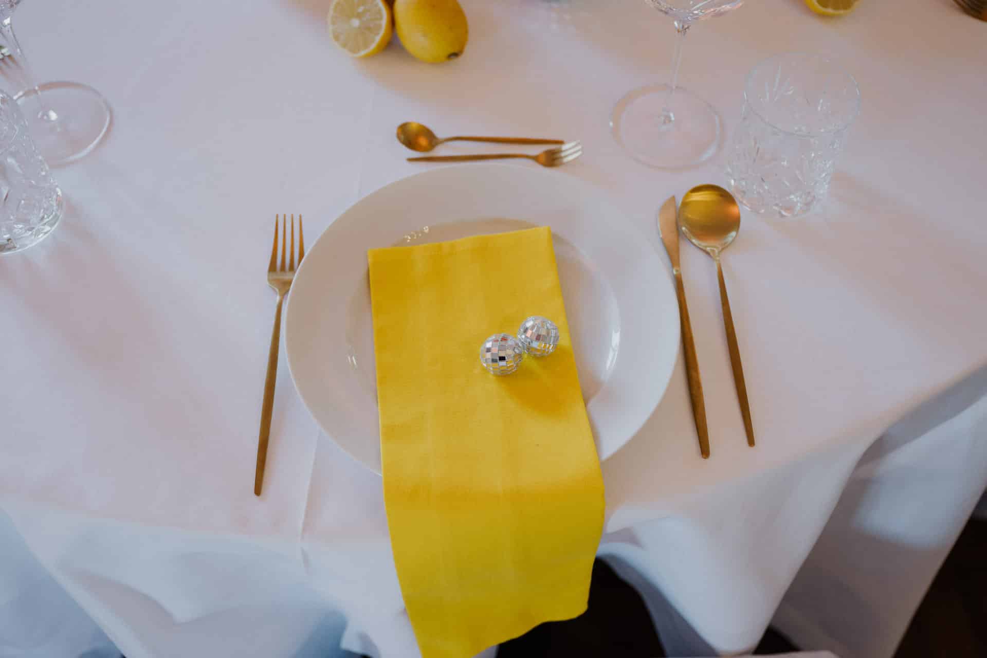 Ein weißer Teller mit einer leuchtend gelben Serviette und zwei kleinen silbernen Kugeln darauf, gedeckt auf einem weißen Tischtuch mit goldenem Besteck und Gläsern. Im Hintergrund sind teilweise Zitronen zu sehen.