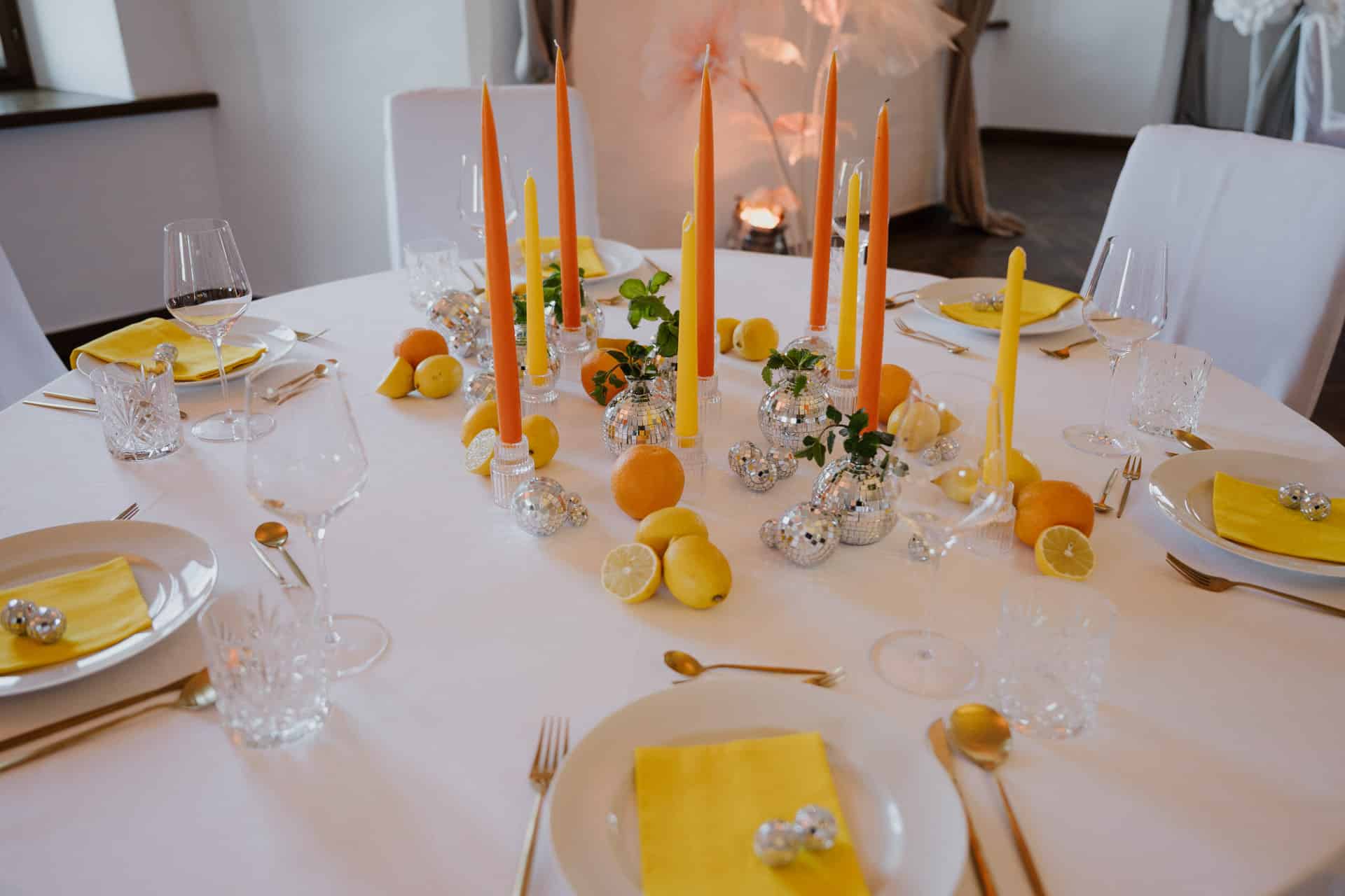 Ein runder weißer Tisch ist mit gelben Servietten, goldenem Besteck, hohen orangefarbenen Kerzen, gläsernen Kerzenhaltern, Zitrusfrüchten, kleinen silbernen Ornamenten und klaren Gläsern für eine Mahlzeit gedeckt.
