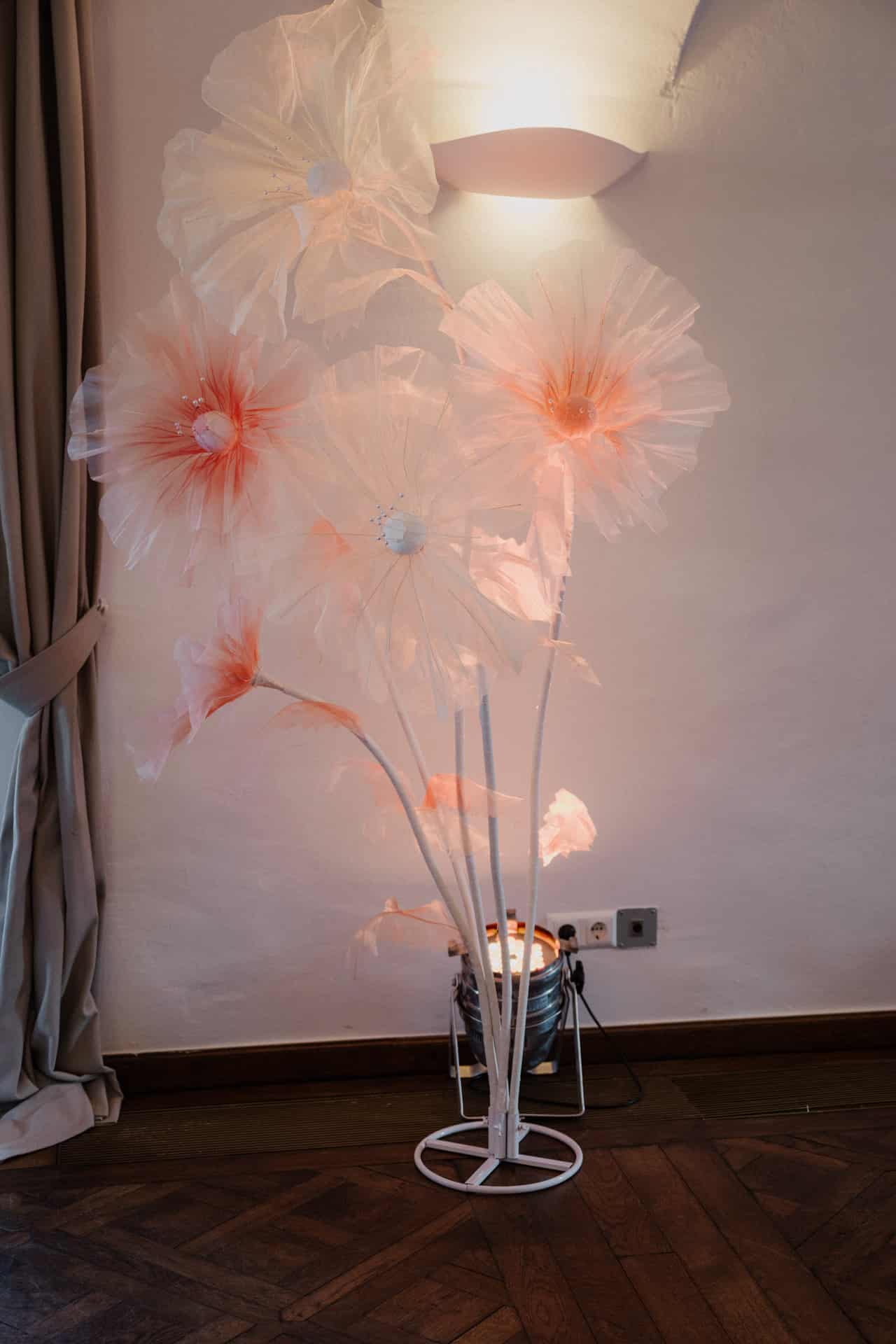 Große, zarte Kunstblumen mit durchscheinenden weißen und rosafarbenen Blütenblättern sind auf hohen Stielen arrangiert und werden von einer sanften Wandleuchte in einem gemütlichen Raum mit Holzfußboden und hellen Vorhängen beleuchtet.