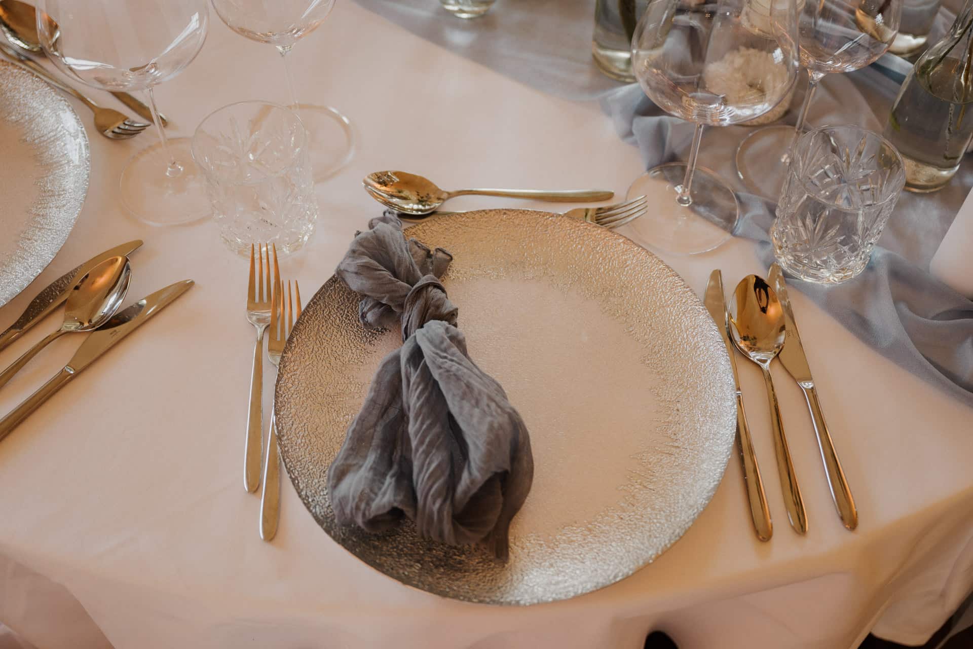 Ein formelles Gedeck auf einem weißen Tischtuch mit einem strukturierten Silberteller, einer grauen geknoteten Serviette, mehreren Weingläsern, einem Kristallwasserglas und poliertem Silberbesteck, das ordentlich auf beiden Seiten des Tellers angeordnet ist.