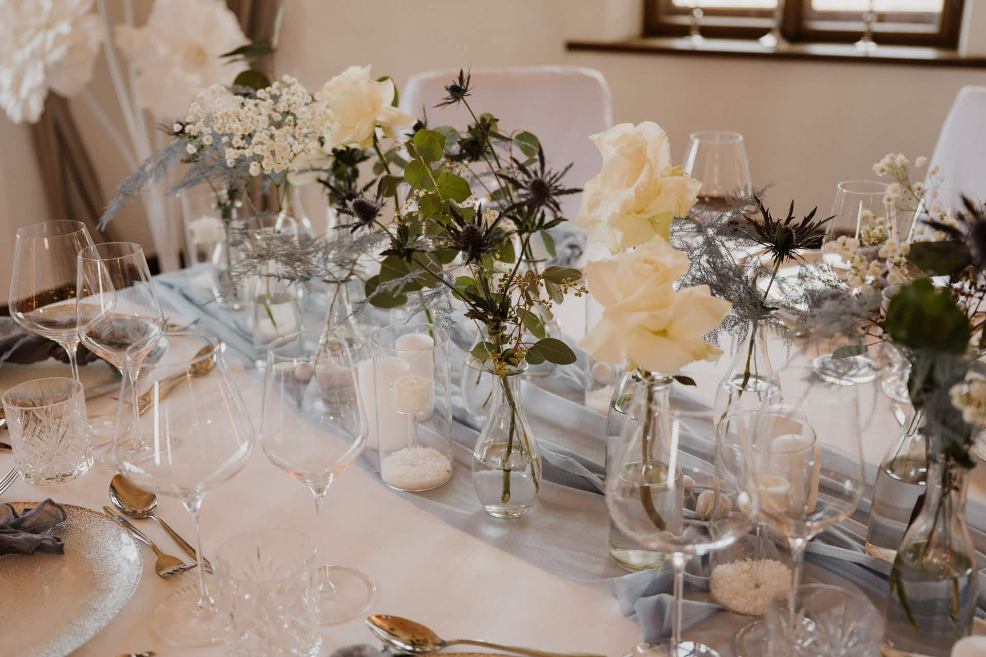 Elegante Tischdekoration mit weißen Rosen, Grünzeug und zarten Blumen in Glasvasen. Weicher blauer Stoff verläuft in der Mitte, umgeben von Glaswaren, Silberbesteck und Kerzen, die eine raffinierte und romantische Atmosphäre schaffen.