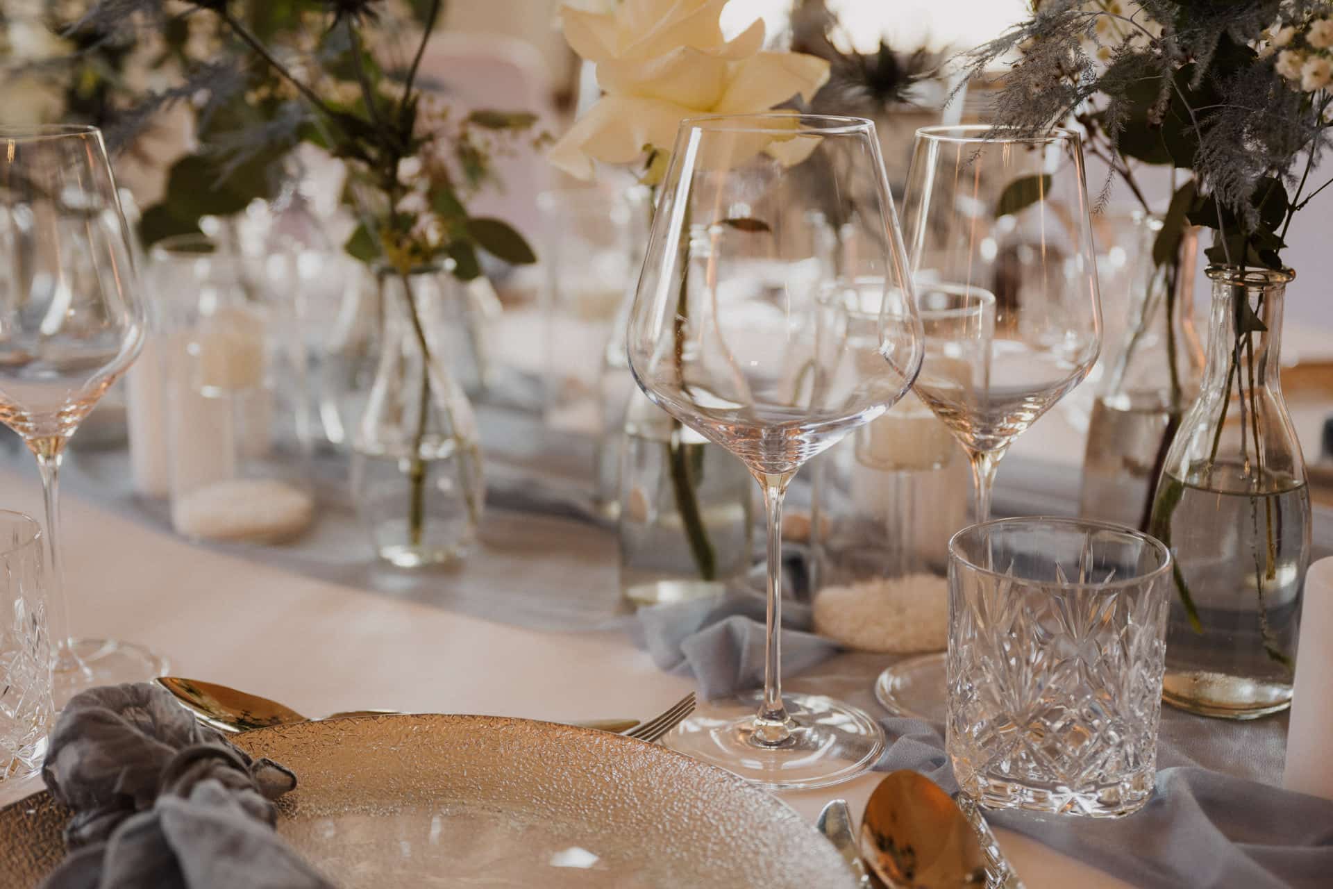 Elegant gedeckter Tisch mit klaren Weingläsern, Glastellern, Silberbesteck und Kristallwassergläsern. Vasen mit weißen Blumen und Grünzeug setzen einen dekorativen Akzent auf einem weichen, neutralen Tischtuch.