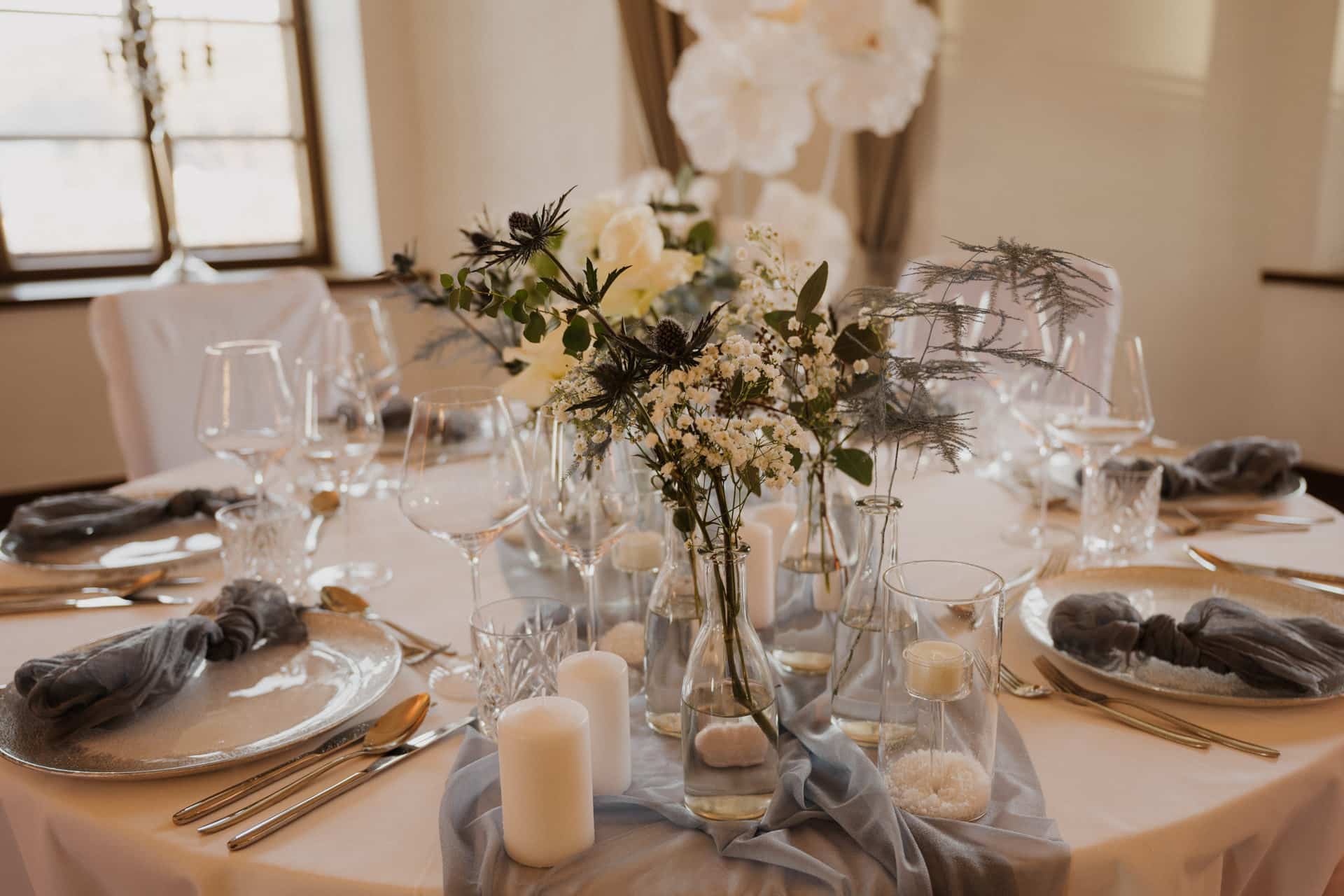 Eleganter runder Tisch für eine formelle Veranstaltung mit weißen Tellern, goldenem Besteck, Weingläsern, weißen Kerzen und zarten Blumensträußen, dekoriert mit sanften blauen Stoffakzenten in einem hellen, luftigen Raum.