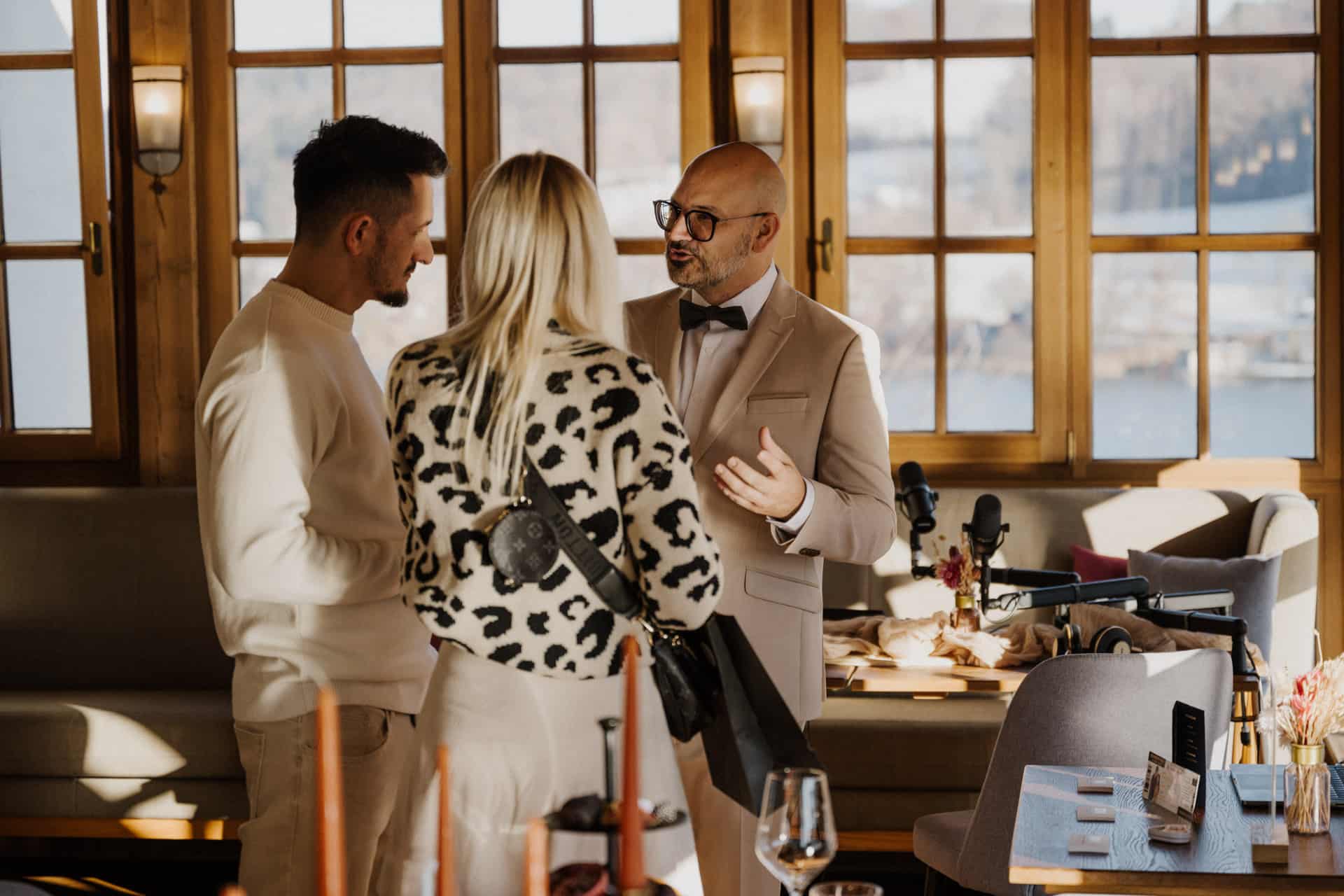 Drei Personen unterhalten sich in einem warm beleuchteten Restaurant an großen Fenstern; ein Mann in einem beigen Anzug gestikuliert, während er mit einem Mann in Weiß und einer Frau in einer Jacke mit Leopardenmuster spricht.