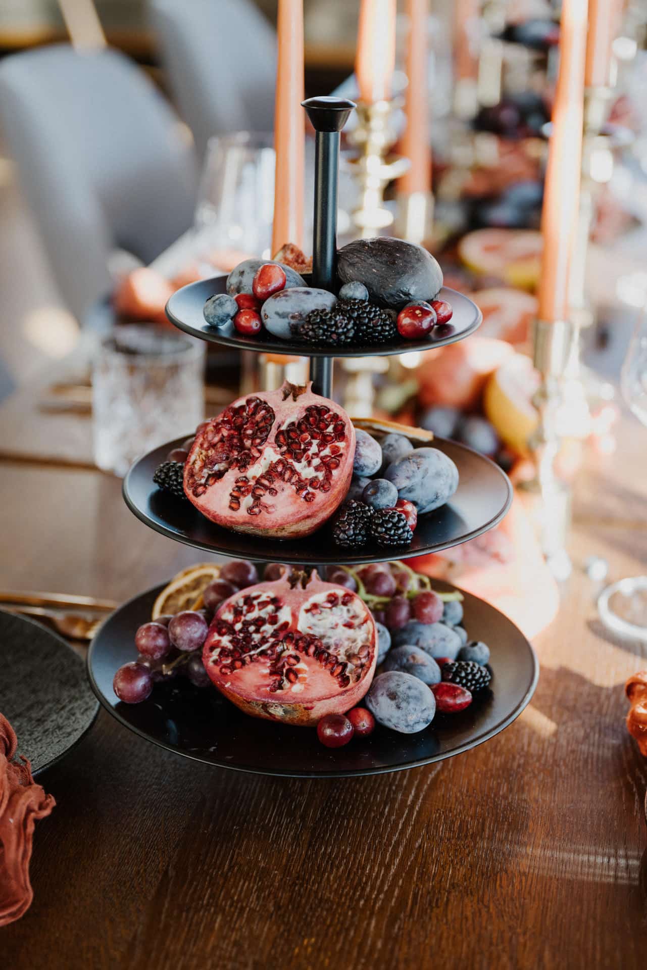 Ein dreistöckiger schwarzer Ständer mit Granatapfelhälften, Pflaumen, Trauben, Brombeeren und Feigen auf einem Holztisch, der für eine Mahlzeit gedeckt ist, mit Kerzen und Glaswaren im Hintergrund.
