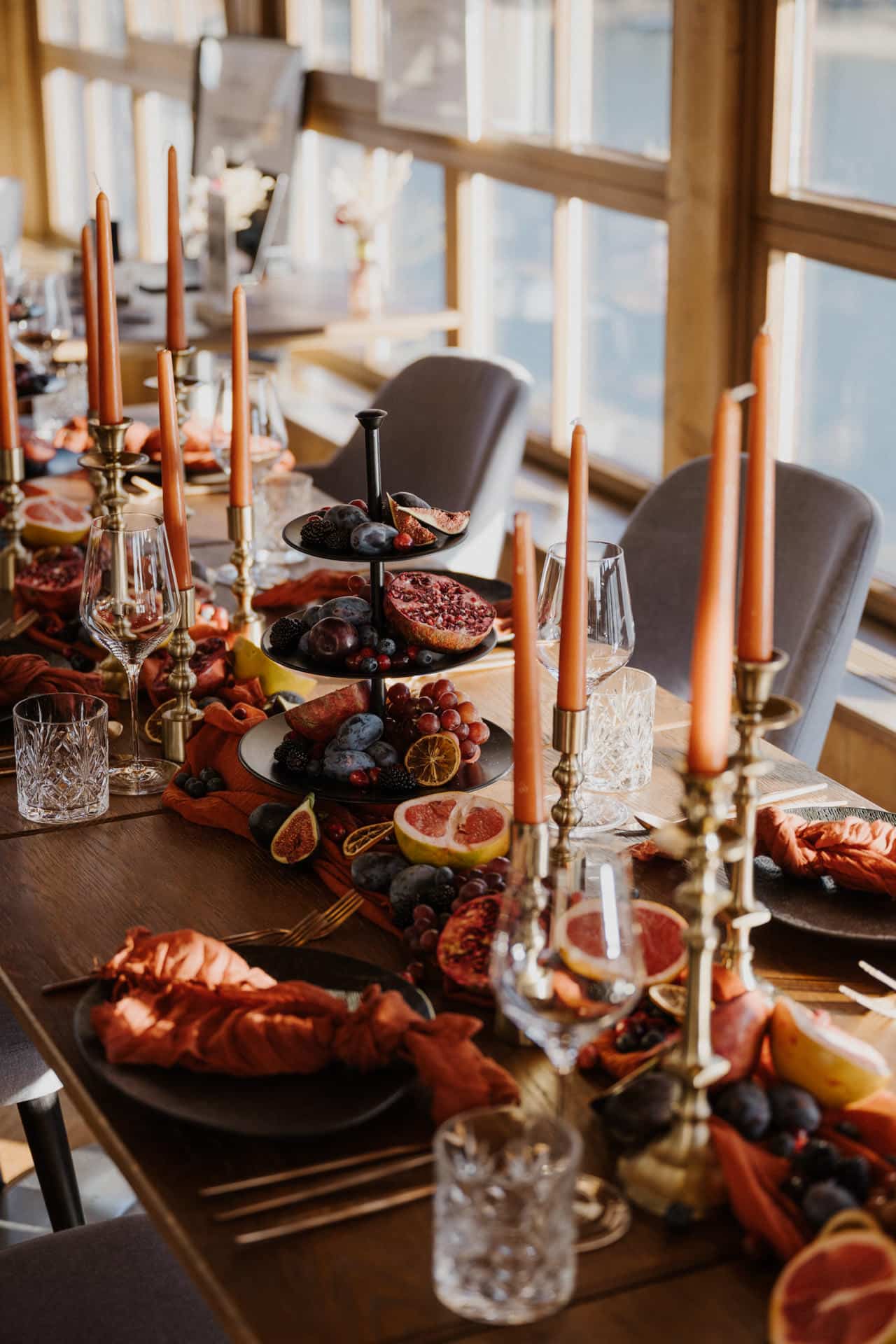 Ein wunderschön gedeckter Esstisch mit orangefarbenen Kerzen, gestapelten Tabletts mit Trauben, Feigen und Granatäpfeln, Gläsern und orangefarbenen Servietten, die durch große Fenster in warmes Sonnenlicht getaucht sind.