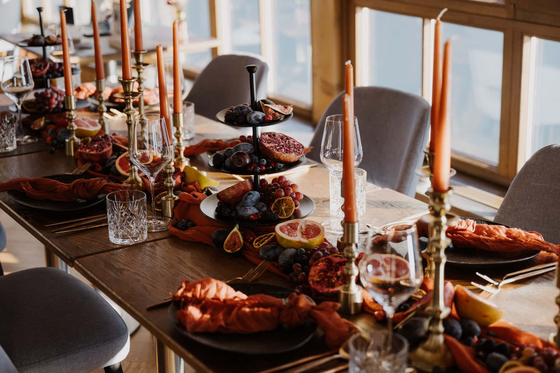 Ein hölzerner Esstisch, gedeckt für eine Mahlzeit mit orangefarbenen Kerzen, Obsttellern mit Trauben, Feigen und Granatäpfeln, Gläsern, Tellern mit orangefarbenen Servietten und goldenem Besteck, im Sonnenlicht durch große Fenster.