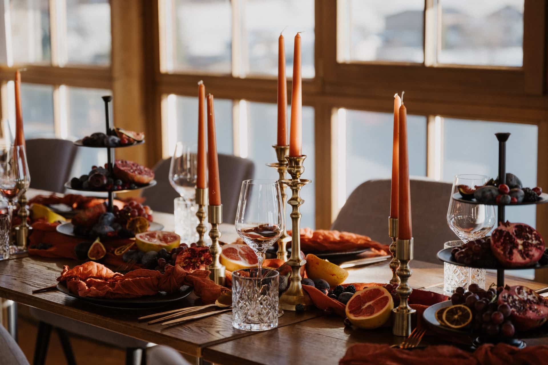 Ein hölzerner Esstisch, der für eine Mahlzeit gedeckt ist, dekoriert mit orangefarbenen Kerzen in Messinghaltern, Weingläsern und Schalen, die mit Früchten wie Trauben, Granatäpfeln und Zitrusfrüchten gefüllt sind. Warmes Sonnenlicht strömt durch große Fenster.