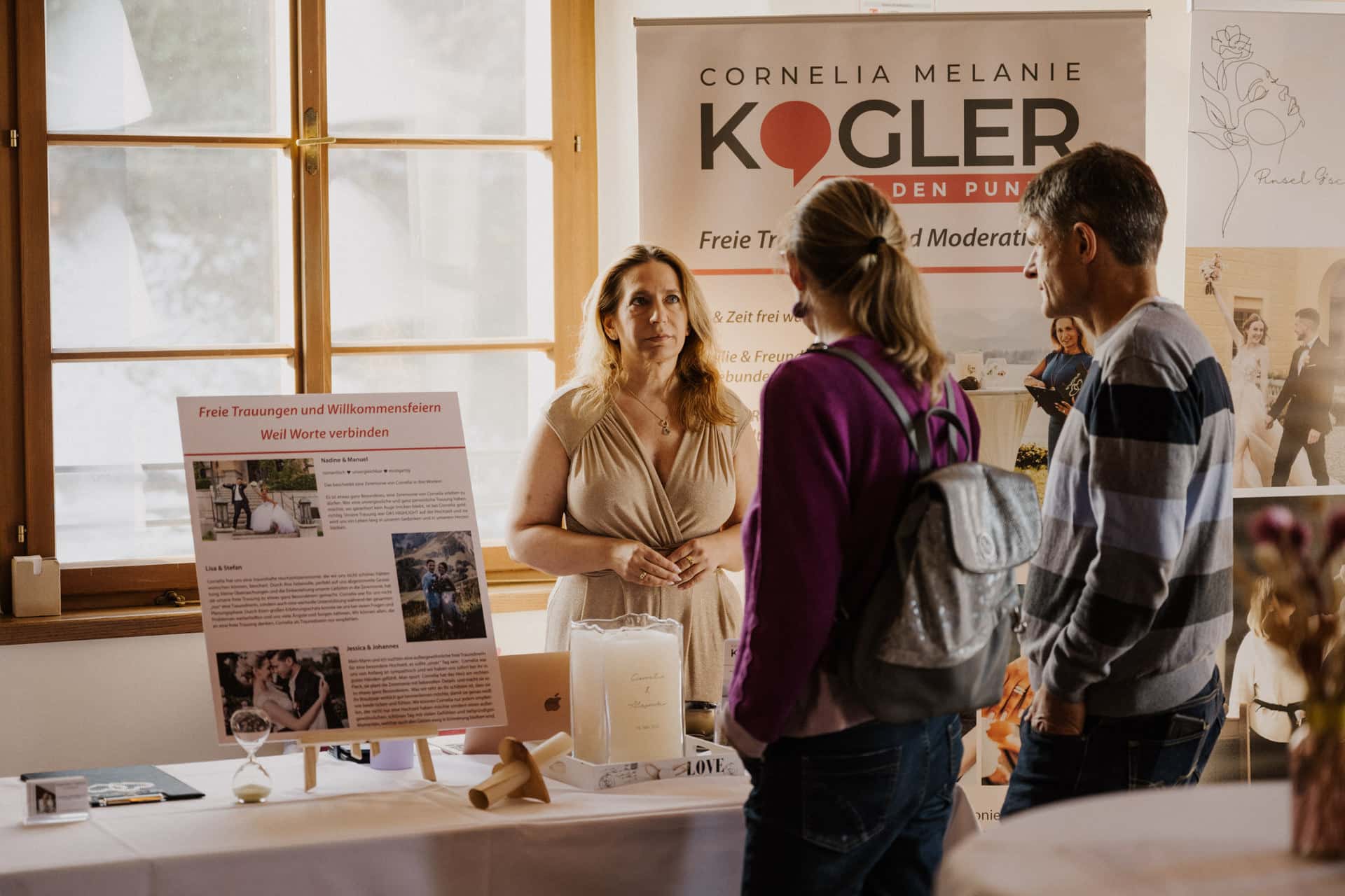 Eine Frau steht bei einer Veranstaltung hinter einem Tisch und spricht mit zwei Personen. Auf dem Tisch befinden sich Informationsdisplays, eine Kerze und Broschüren. Im Hintergrund ist ein Transparent mit "Cornelia Melanie Kogler" und Hochzeitsfotos zu sehen.