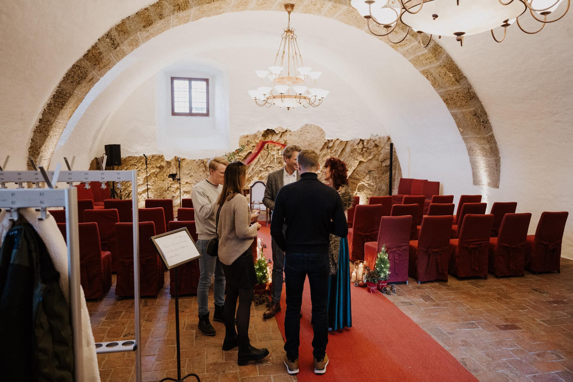 Eine kleine Hochzeitszeremonie in einem rustikalen Raum mit gewölbter Steindecke, roten Stühlen und einem roten Teppich. Vier Personen stehen vor dem Altar, umgeben von festlicher Dekoration und sanfter Beleuchtung.