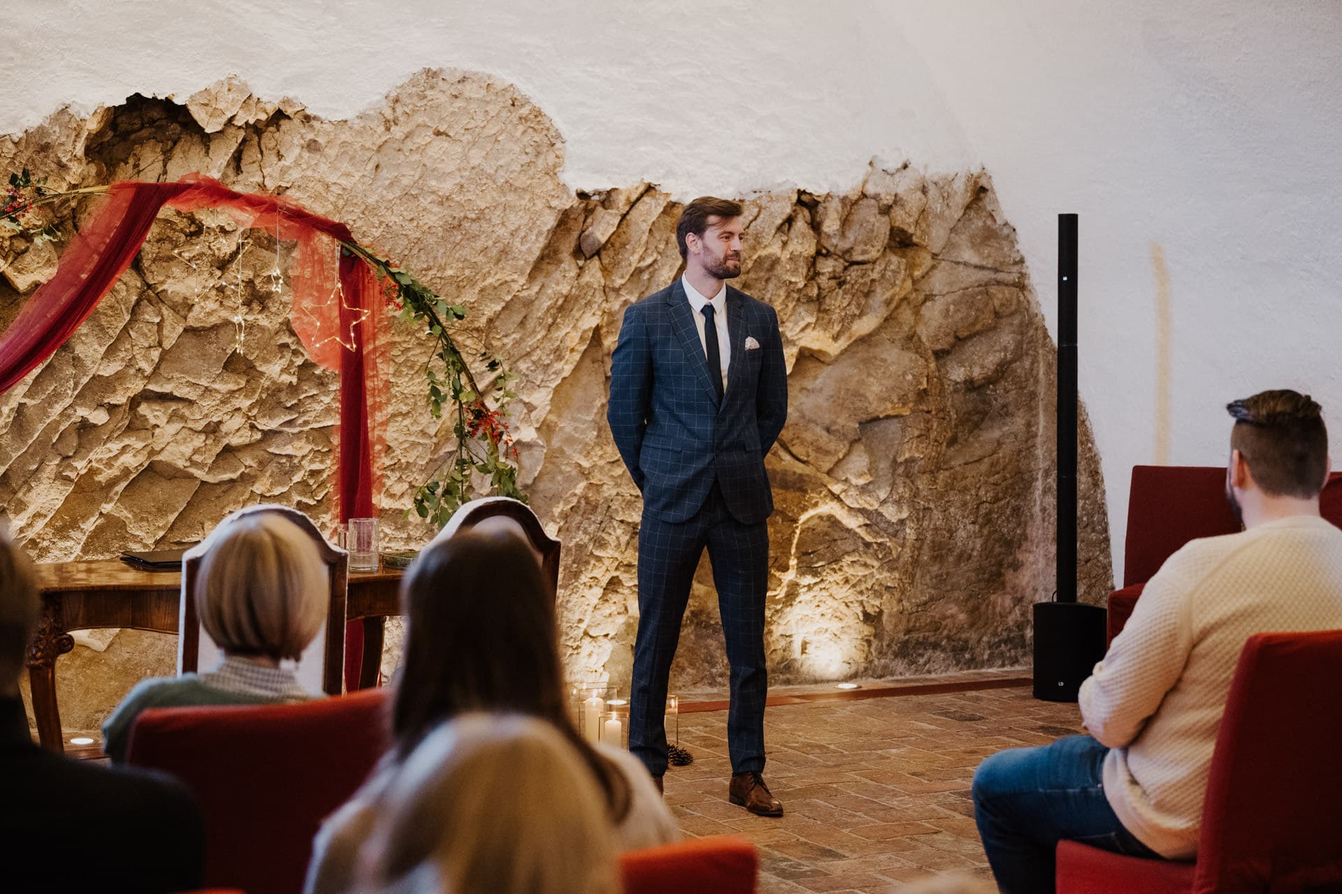 Ein Mann in einem blaukarierten Anzug steht vor einem kleinen sitzenden Publikum in einem rustikalen Raum mit einer Steinwand und einem rot verhangenen Bogen und scheint eine Rede zu halten oder eine Veranstaltung zu moderieren.