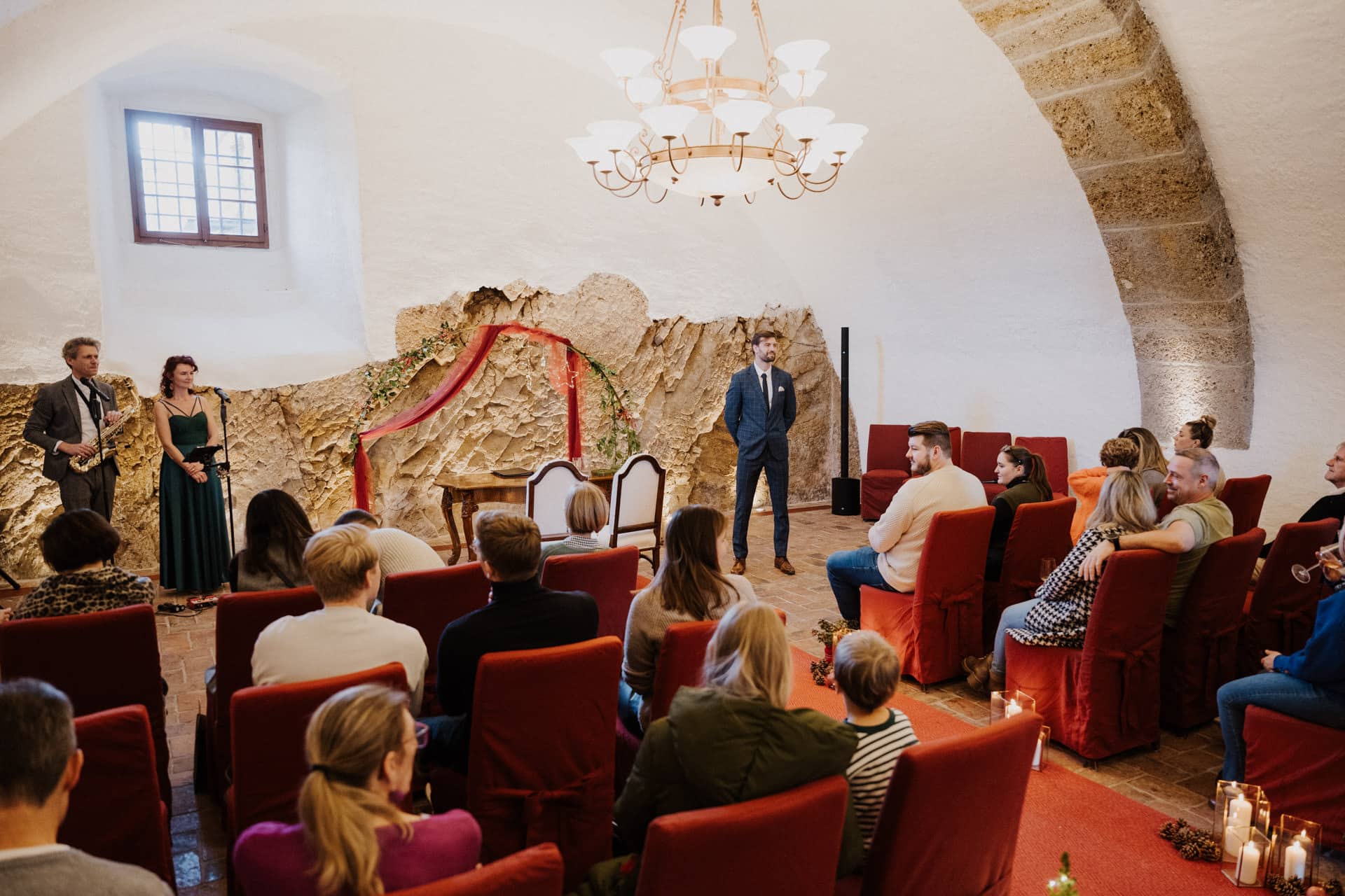 Eine Gruppe von Menschen sitzt auf roten Stühlen vor einer Hochzeitszeremonie in einem rustikalen Raum mit Steinwänden, einer gewölbten Decke und einem Kronleuchter. Drei Personen stehen vorne, in der Nähe eines Tisches und zweier Stühle, umrahmt von Grünzeug.