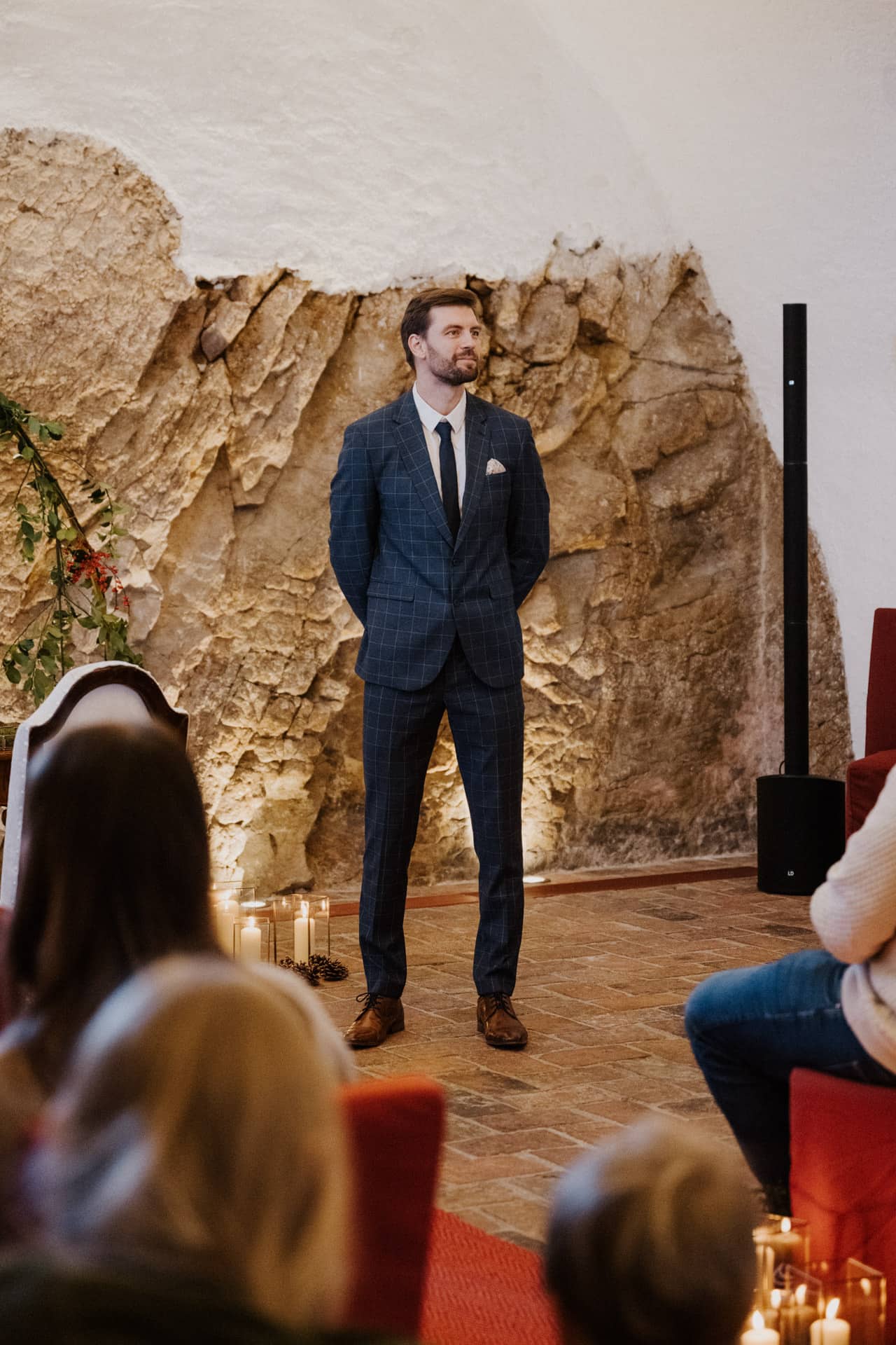 Ein Mann in einem blaukarierten Anzug steht mit den Händen auf dem Rücken in einem rustikalen Raum mit Steinwänden und spricht zu einem vor ihm sitzenden Publikum; der Boden ist mit Kerzen geschmückt.