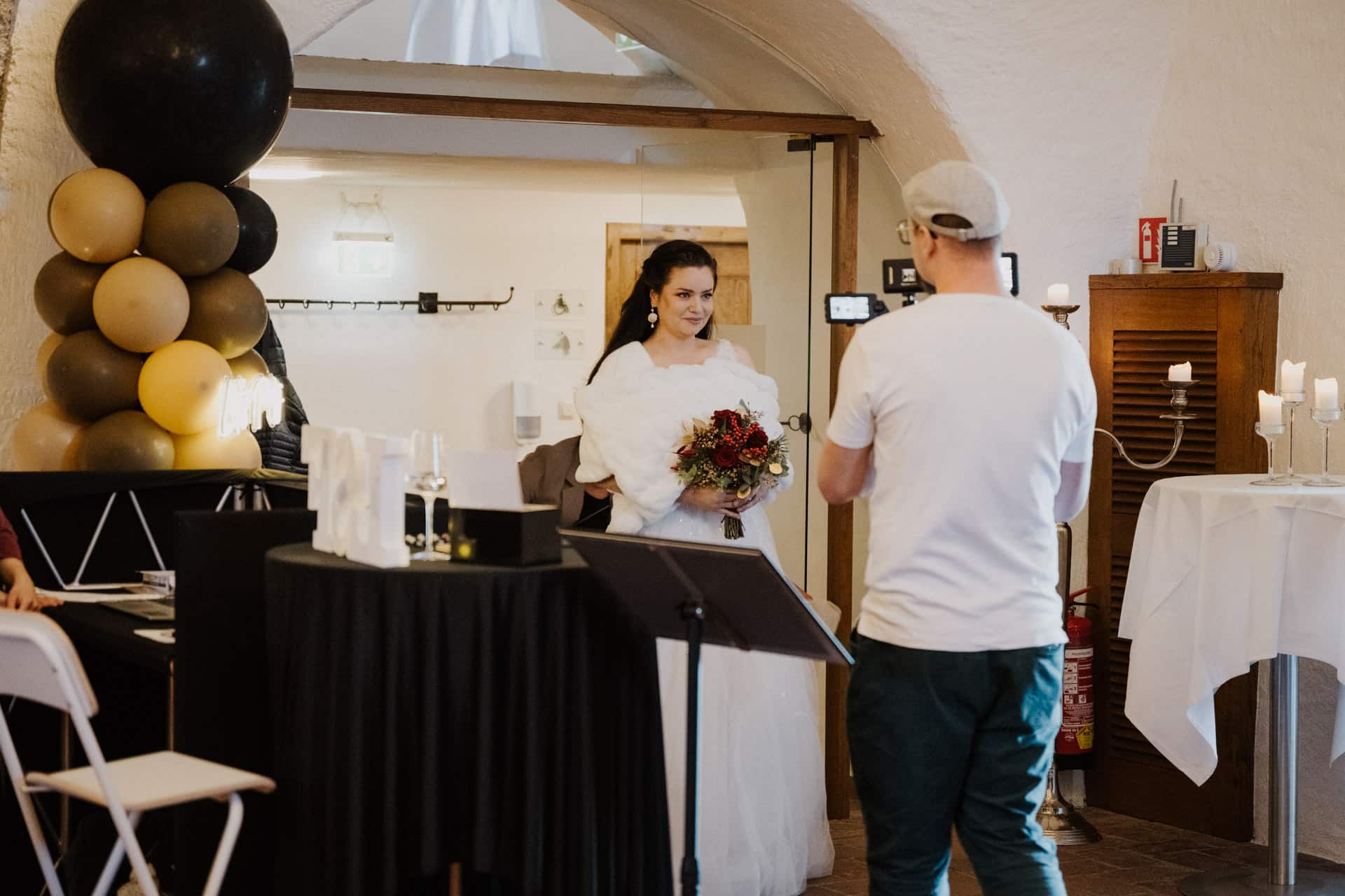Eine Braut in einem weißen Kleid und mit einem Blumenstrauß in der Hand steht in einem Haus und blickt auf eine Person, die sie mit einer Kamera filmt. Luftballons, Kerzen und ein Tisch stehen in der Nähe und schaffen eine festliche Atmosphäre.