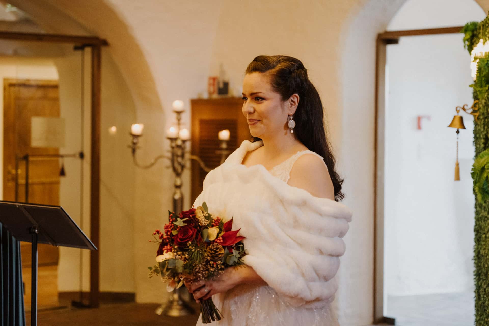 Eine Braut in einem weißen Kleid und einer Pelzstola hält einen Strauß roter und weißer Blumen in der Hand. Sie steht in einem Innenraum in der Nähe eines Kandelabers und Grünzeugs und hat ein sanftes Lächeln im Gesicht.