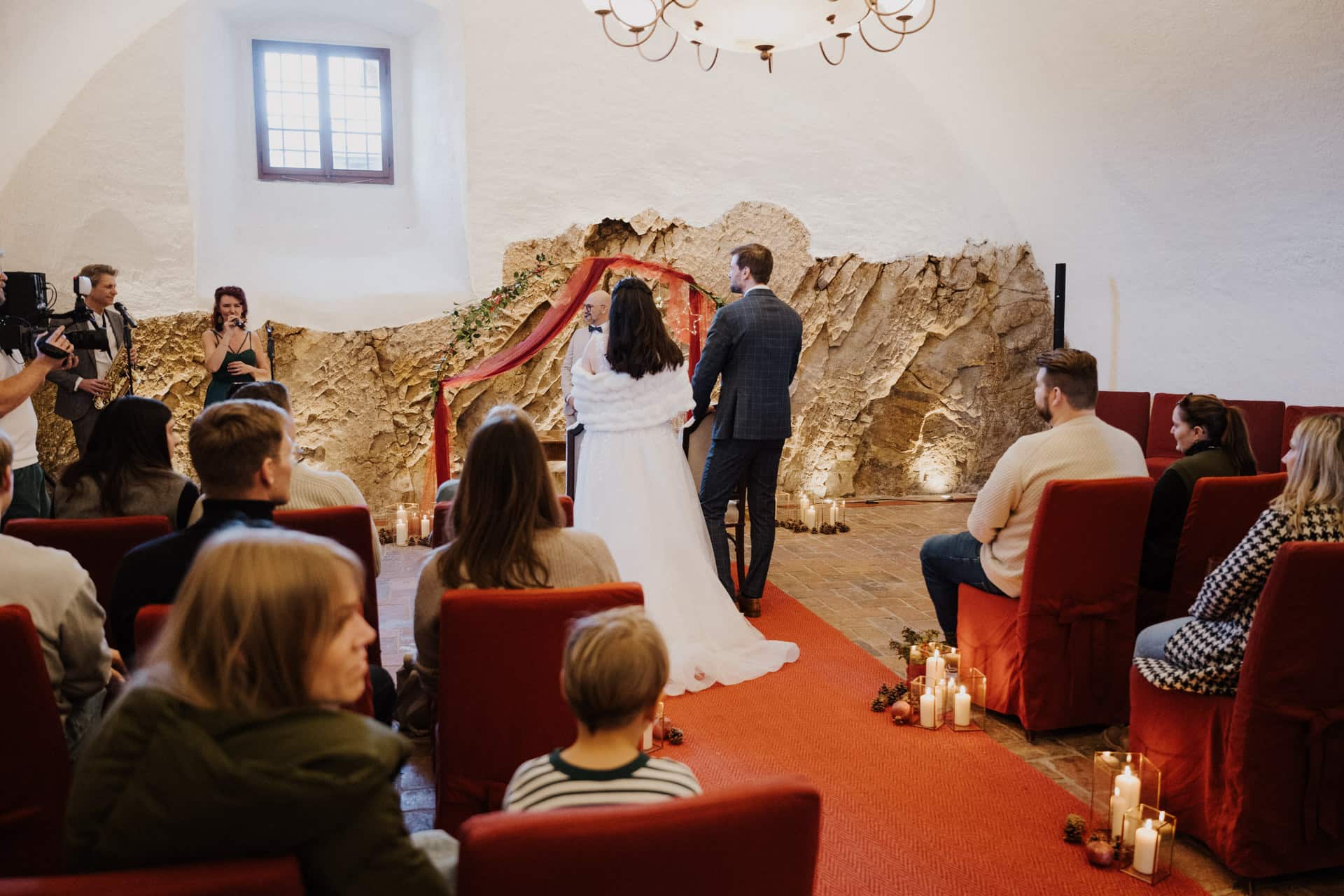 Braut und Bräutigam stehen vor dem Altar in einem kleinen, rustikalen Raum mit Steinwänden, umgeben von sitzenden Gästen. Kerzen säumen den Gang, und ein Fotograf hält den Moment fest.