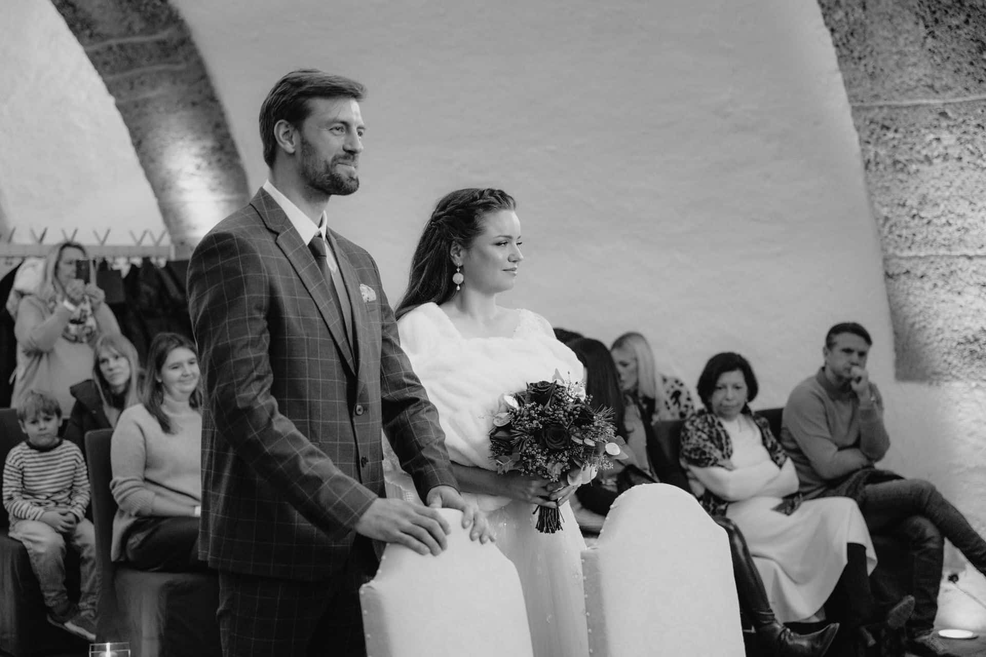 Braut und Bräutigam stehen während einer Hochzeitszeremonie Seite an Seite im vorderen Teil eines Raumes, umgeben von sitzenden Gästen. Der Bräutigam trägt einen karierten Anzug, und die Braut hält einen Blumenstrauß. Das Foto ist in schwarz-weiß gehalten.