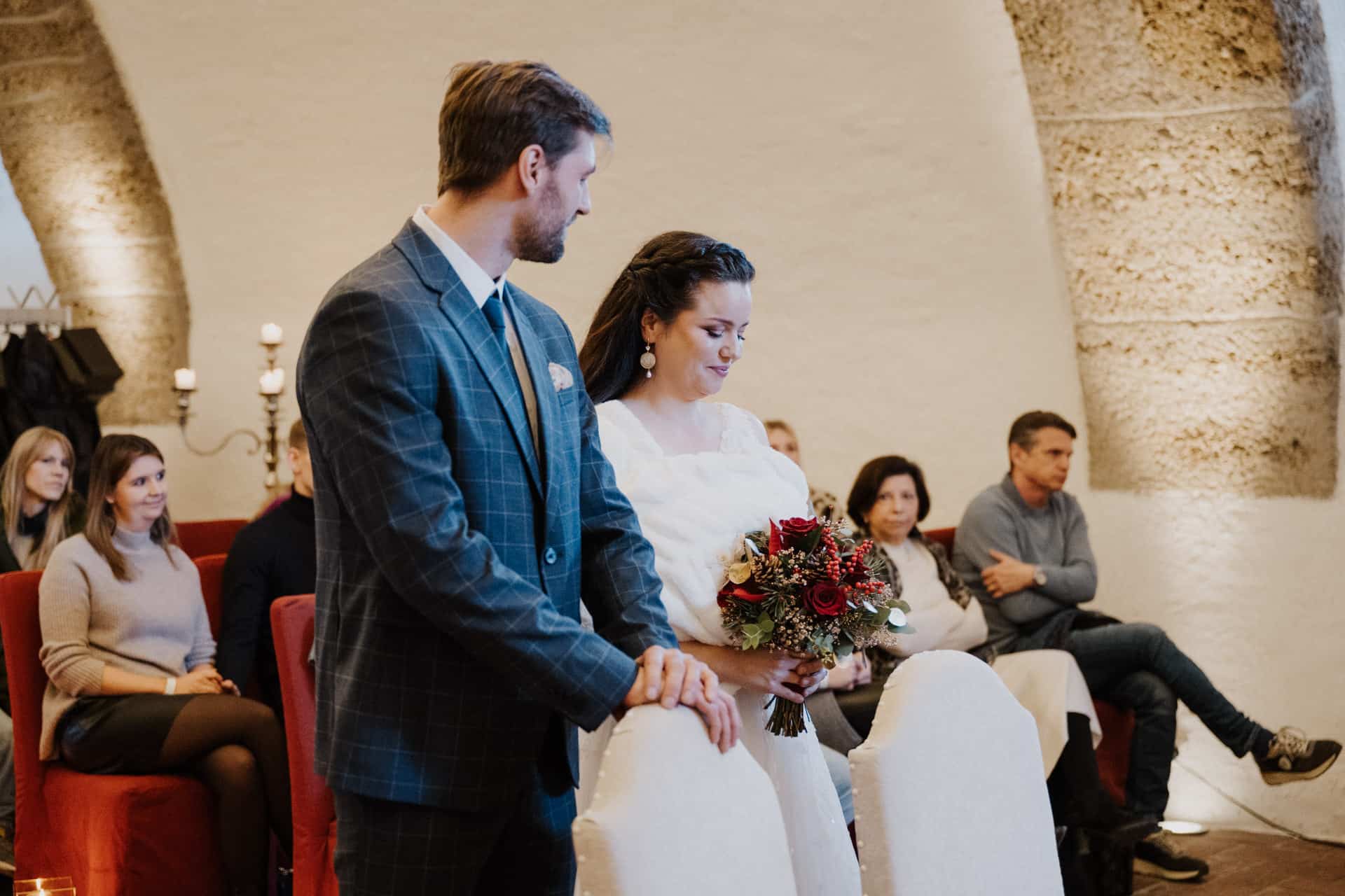 Eine Braut, die einen Blumenstrauß hält, und ein Bräutigam stehen während einer Hochzeitszeremonie in einem Innenraum zusammen, während die Gäste im Hintergrund aufmerksam zusehen.