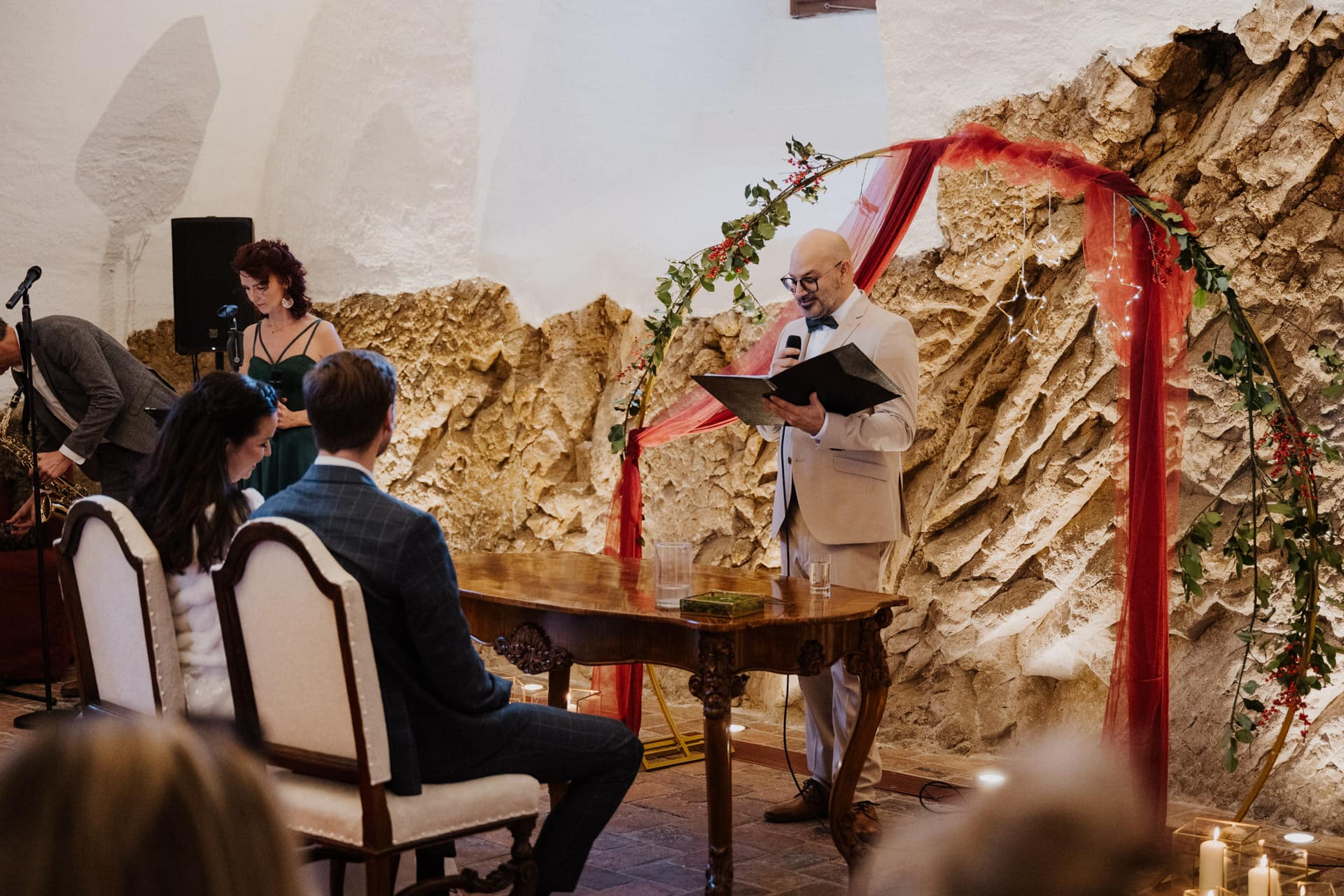 Ein Trauredner im hellen Anzug liest an einem Holztisch mit Blick auf ein sitzendes Paar aus einer Mappe vor. Die Zeremonie findet drinnen statt, mit rustikalen Steinwänden, roten Vorhängen, Grünzeug und Gästen, die zusehen.
