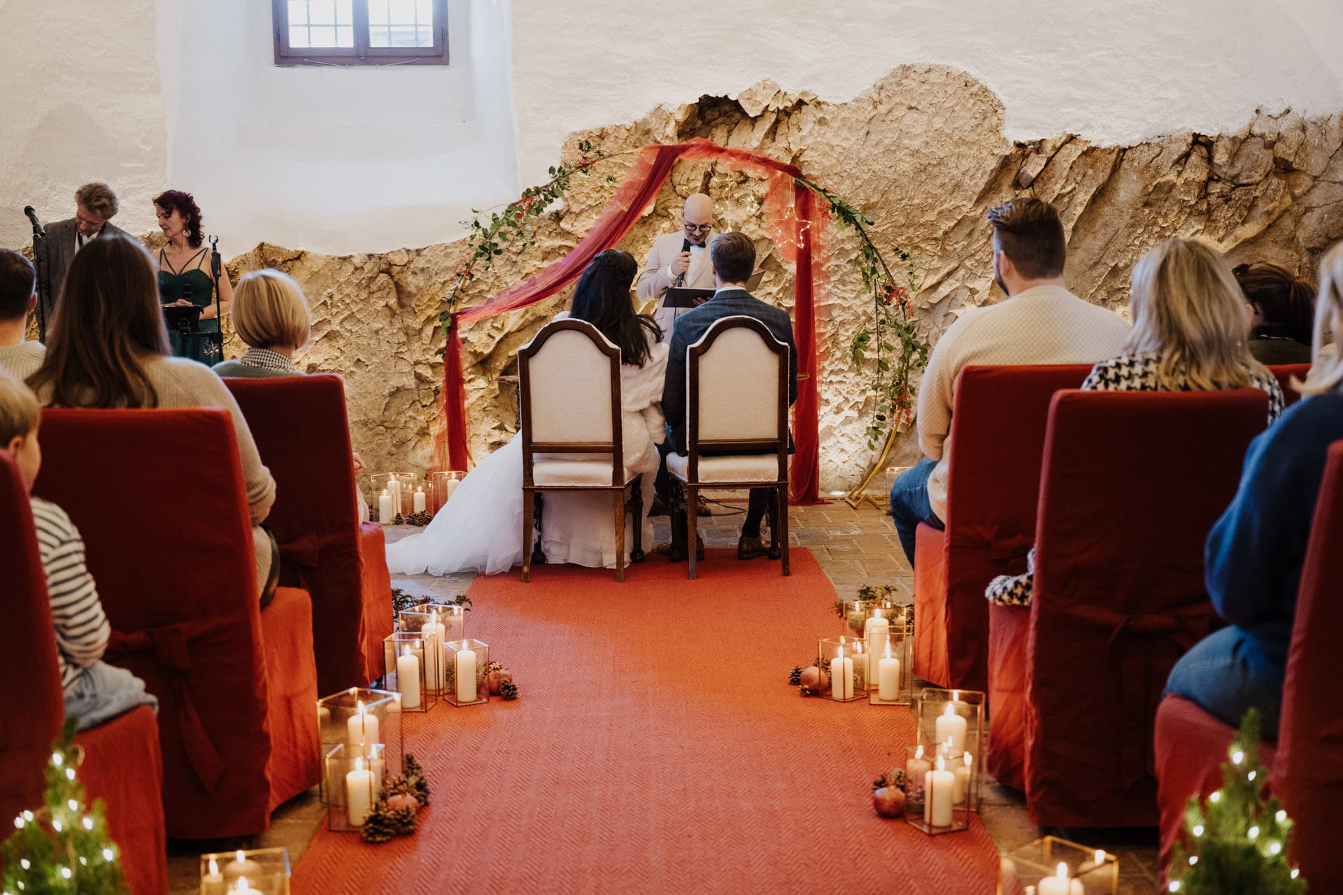 Braut und Bräutigam sitzen während einer Hochzeitszeremonie einem Geistlichen gegenüber. Die Gäste sitzen auf rot bezogenen Stühlen, und der Gang ist mit Kerzen und Grünzeug gesäumt. Der Veranstaltungsort verfügt über eine Natursteinwand und eine warme, intime Beleuchtung.