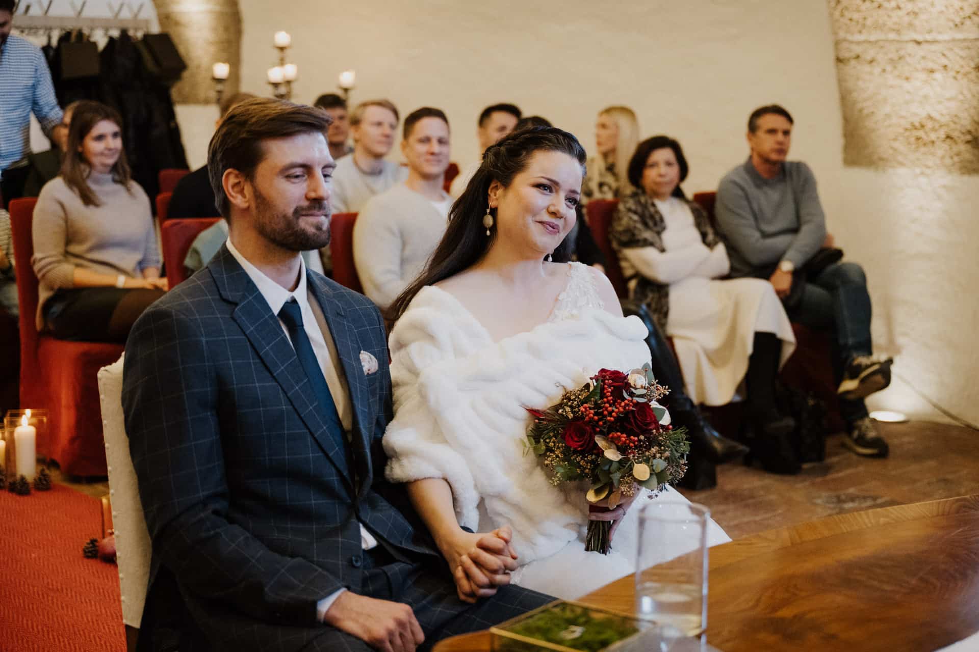 Eine Braut und ein Bräutigam sitzen nebeneinander an einem Holztisch und halten sich während einer Hochzeitszeremonie an den Händen. Die Braut trägt ein weißes Kleid und hält einen Blumenstrauß, während die Gäste hinter ihnen in einem warm beleuchteten Raum sitzen.