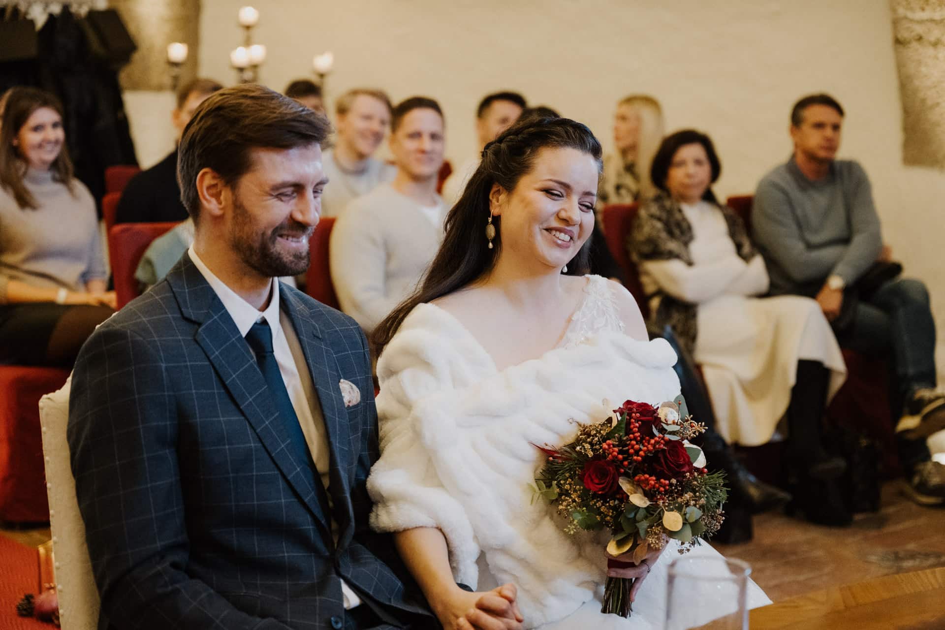 Eine Braut in einem weißen Kleid mit einem Strauß roter und weißer Blumen sitzt neben einem Bräutigam in einem karierten Anzug. Sie lächeln, während die Gäste, die hinter ihnen sitzen, die Zeremonie in einem warm beleuchteten Raum verfolgen.