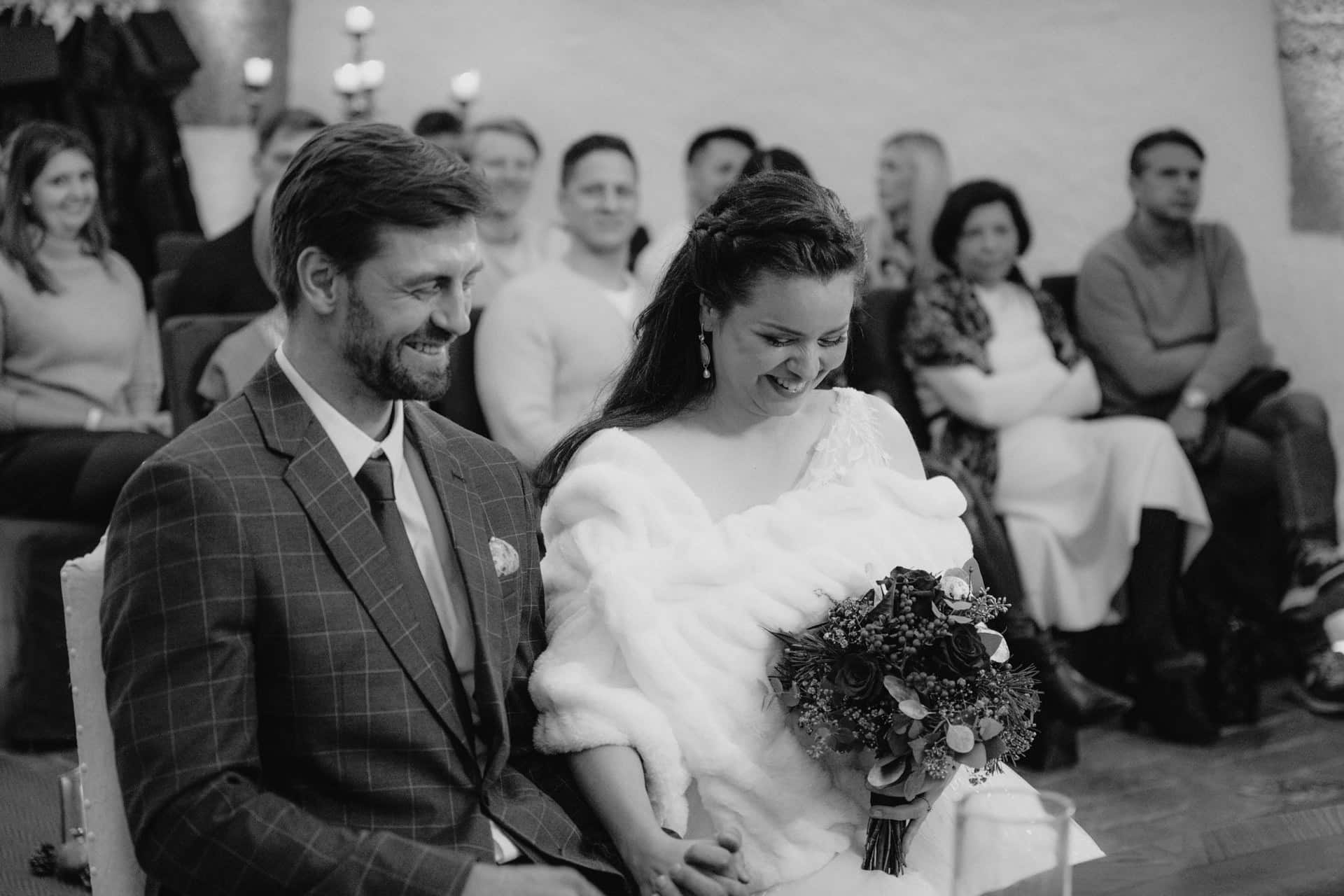 Eine Braut und ein Bräutigam sitzen während einer Hochzeitszeremonie lächelnd zusammen. Die Braut hält einen Blumenstrauß und trägt ein weißes Tuch, während der Bräutigam einen karierten Anzug trägt. Die Gäste sitzen hinter ihnen und schauen aufmerksam zu.