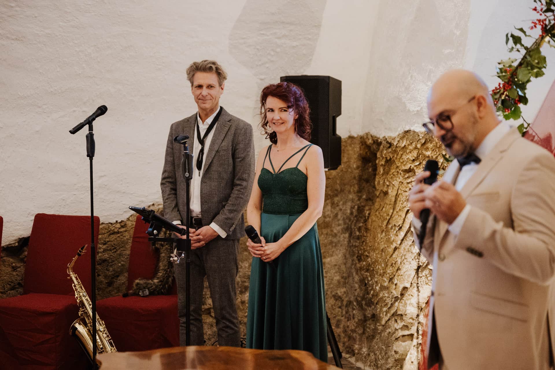 Ein Mann und eine Frau stehen nebeneinander und halten Mikrofone, während ein anderer Mann in einem beigen Anzug in ein Mikrofon im Vordergrund spricht. Ein Saxophon steht auf einem Ständer in der Nähe. Sie befinden sich in einem Raum mit weißen Wänden und roten Stühlen.
