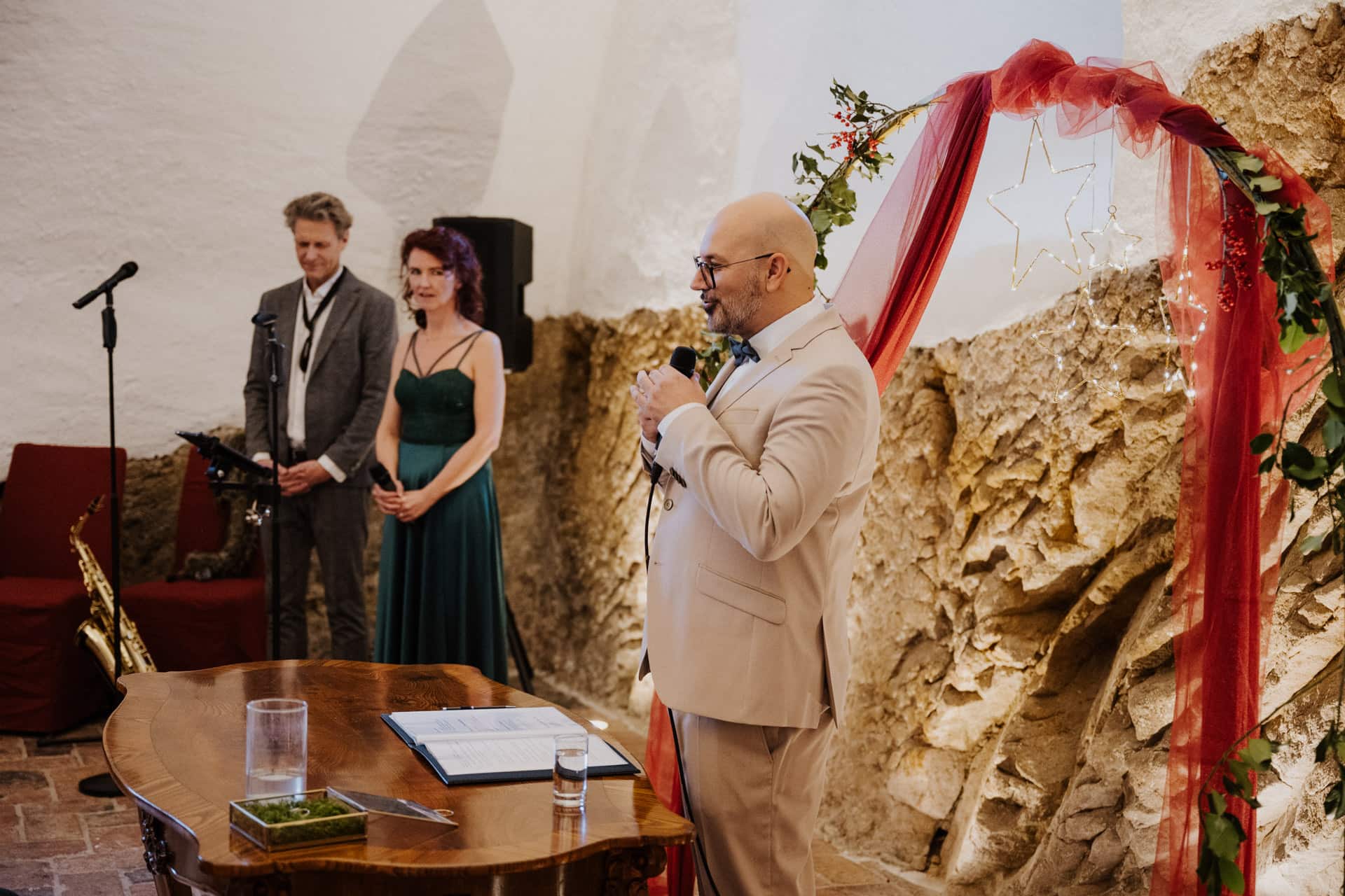 Ein Mann in einem hellen Anzug spricht bei einer Hochzeitszeremonie neben einem geschmückten Bogen in ein Mikrofon. Eine Frau in einem grünen Kleid und ein Mann in einem grauen Anzug stehen daneben. Im Vordergrund steht ein Tisch mit Papieren und Gläsern.